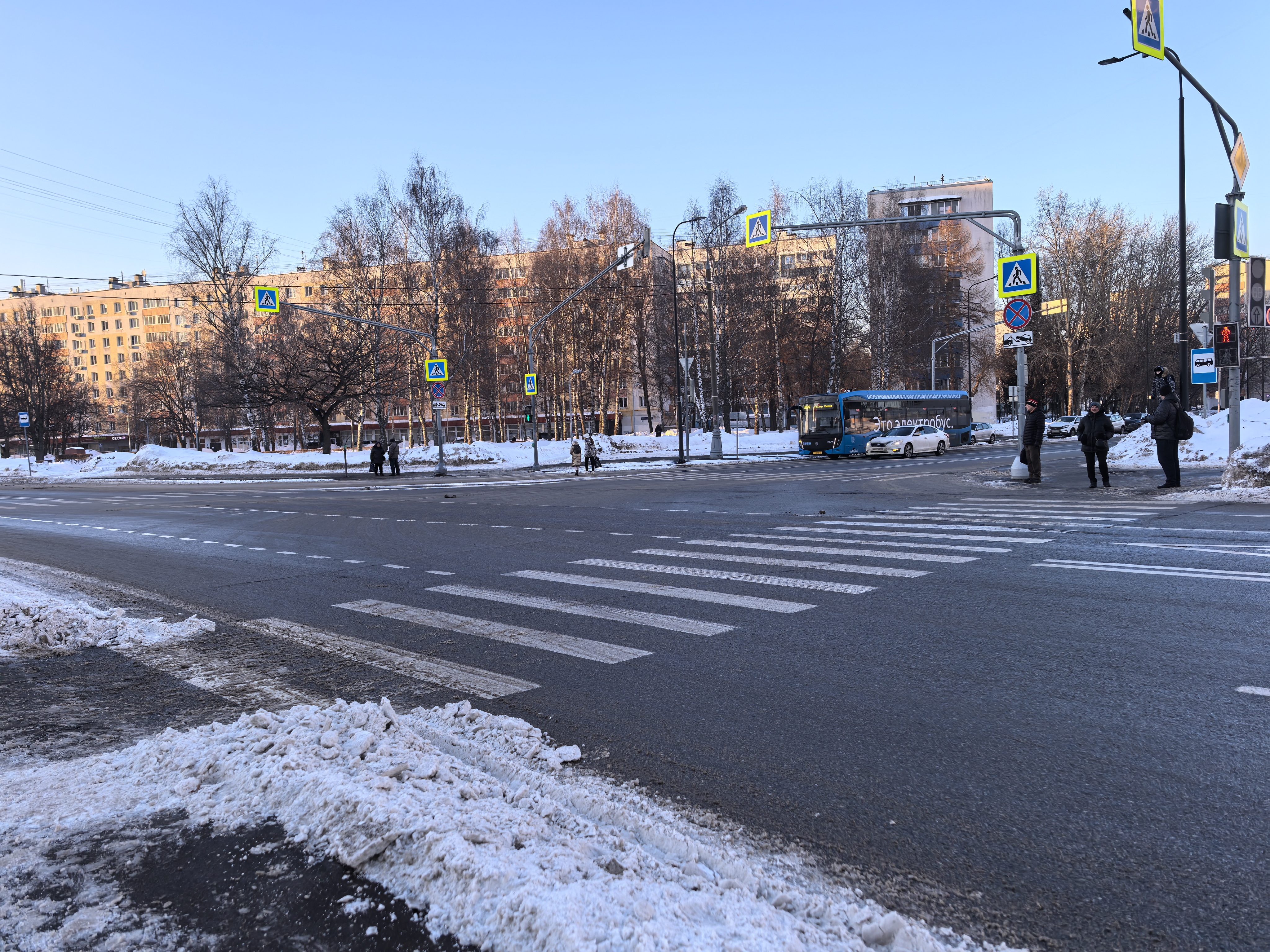 Широкая городская улица с пешеходным переходом, регулируемым светофором. Видны зимние условия, снег на обочинах, но проезжая часть очищена.