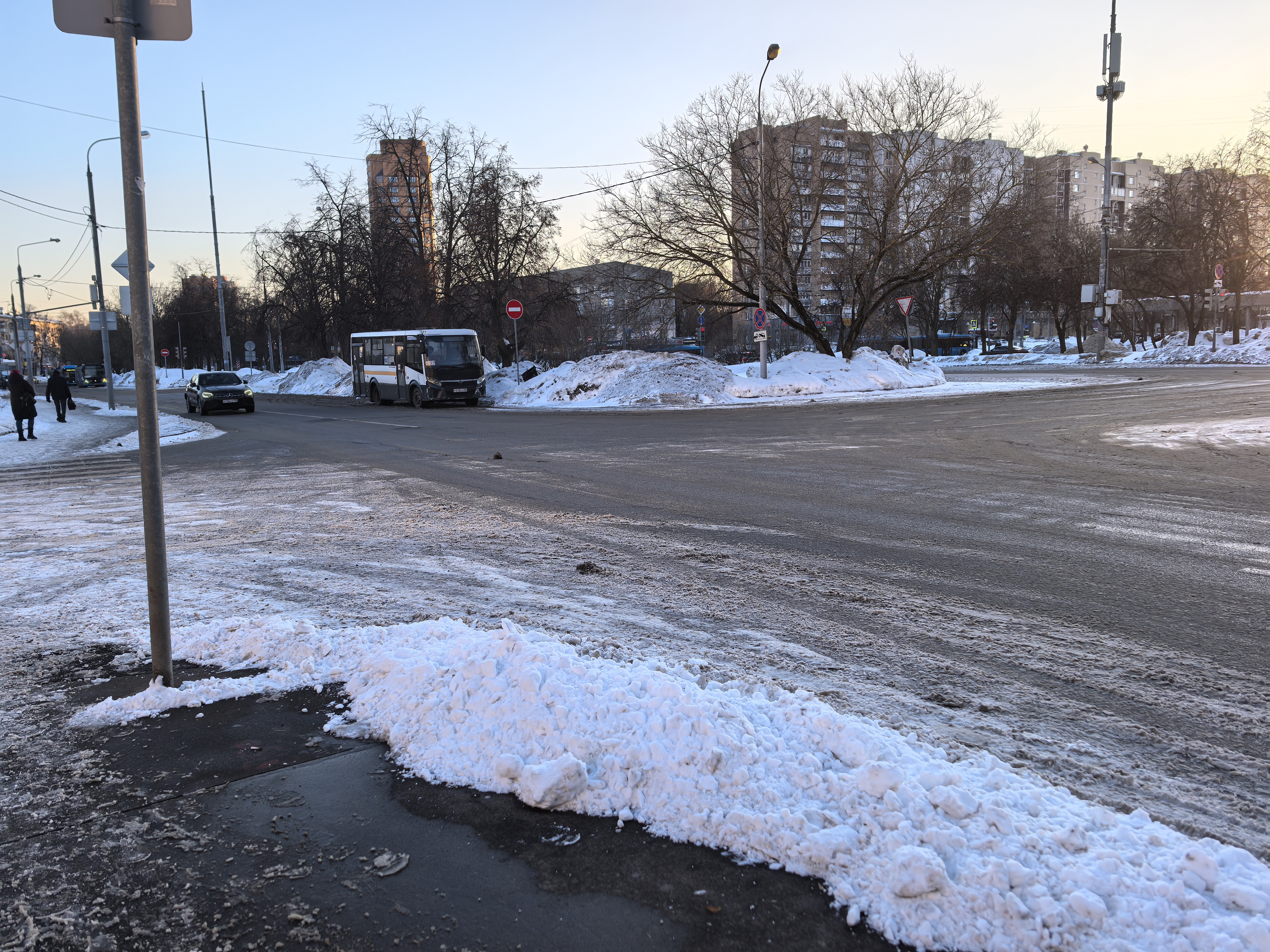 Участок проезжей части без пешеходного перехода, видны тротуары, припаркованный автобус и движущийся автомобиль. Дорога покрыта снегом и льдом, зимний период.