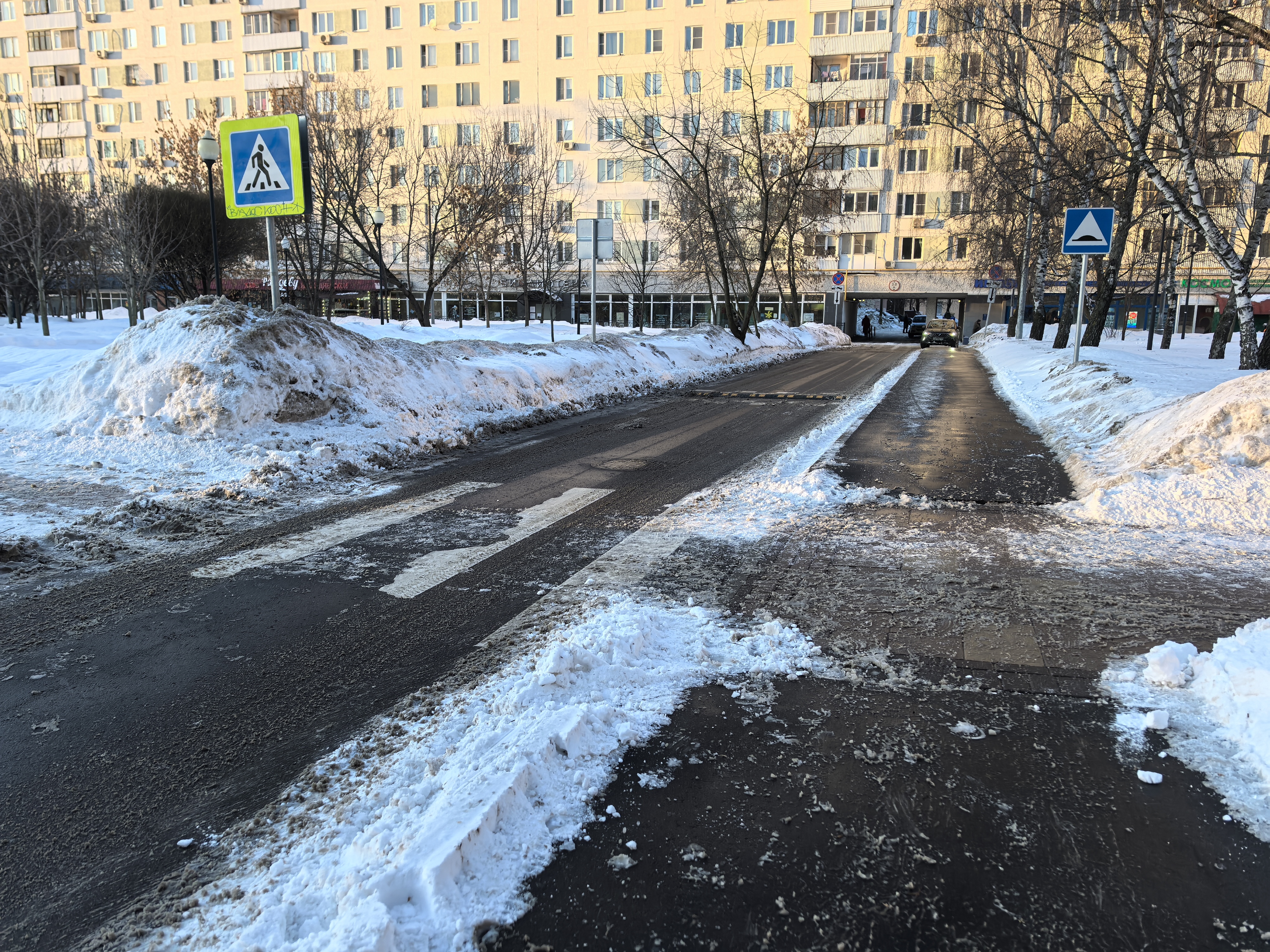 Проезжая часть с пешеходным переходом (зебра) в зимний период, рядом с жилым домом. Дорога частично очищена от снега, но покрыта слежавшимся снегом и наледью. Видны лежачий полицейский и знаки.