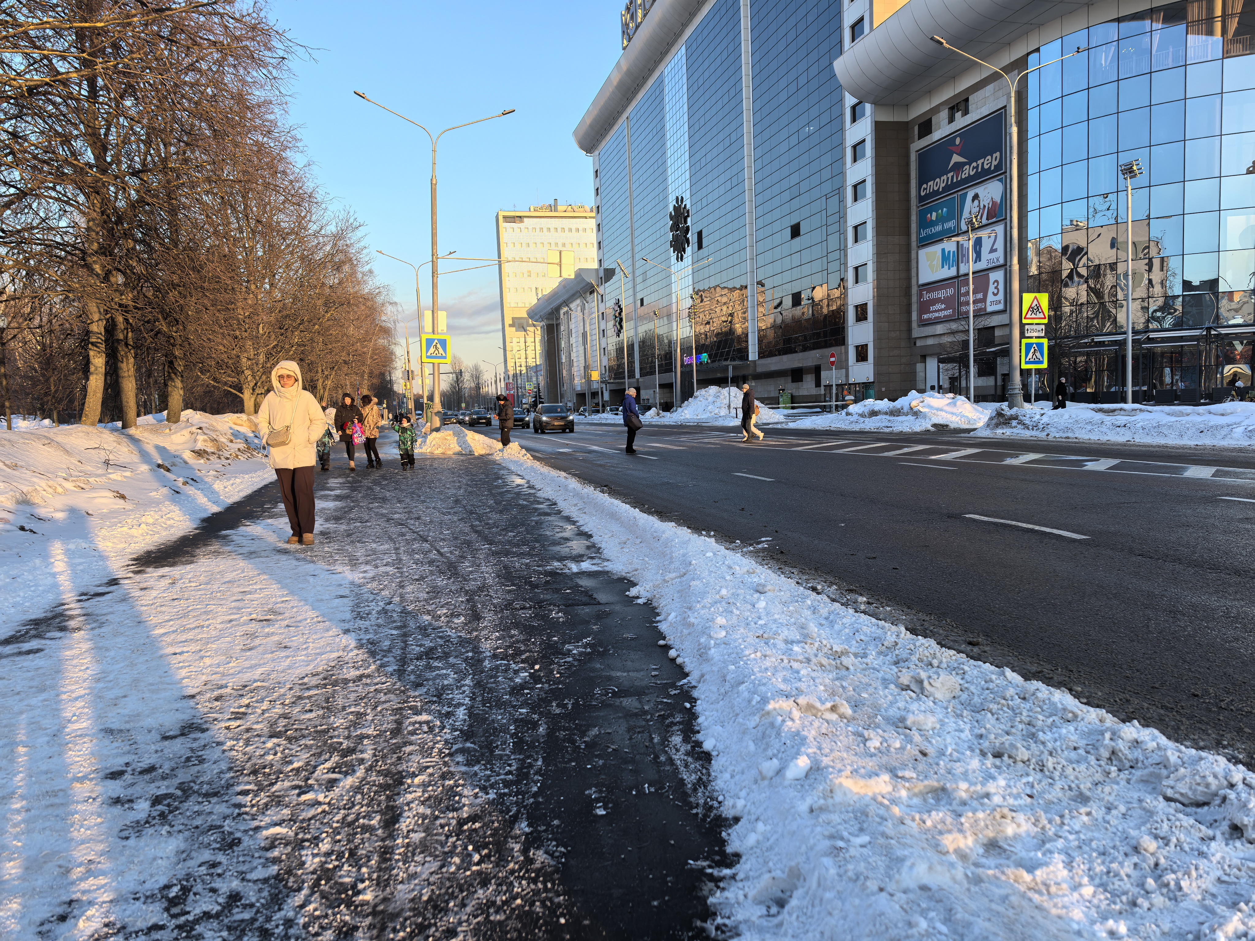 Городская улица с пешеходным переходом, тротуаром и прилегающим парком. Видны пешеходы, включая детей, и проезжающие автомобили.