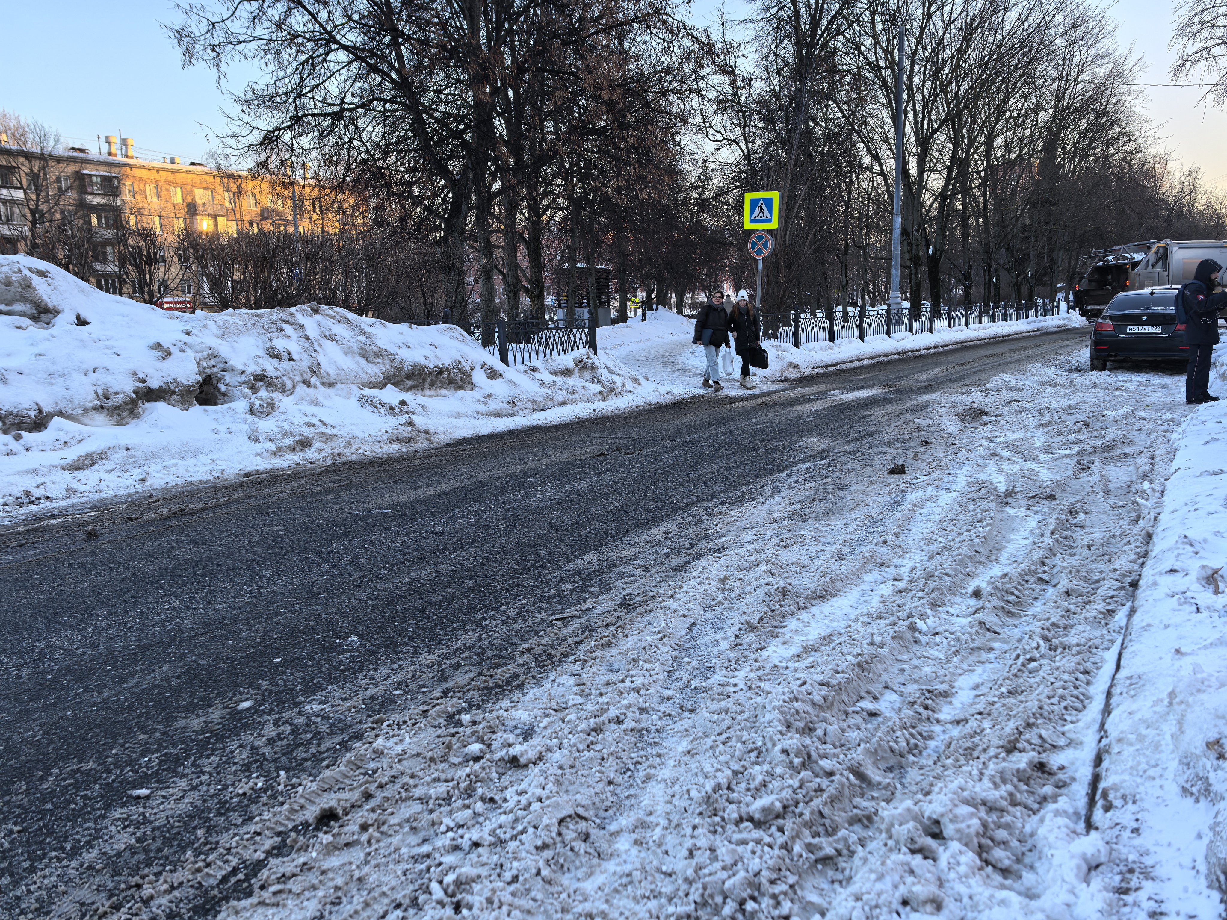 Участок городской дороги без видимой пешеходной разметки (зебры) на проезжей части. Справа установлен знак пешеходного перехода, указывающий на его наличие впереди, но сам переход в кадре отсутствует. Дорога покрыта снегом и слякотью, тротуар частично очи