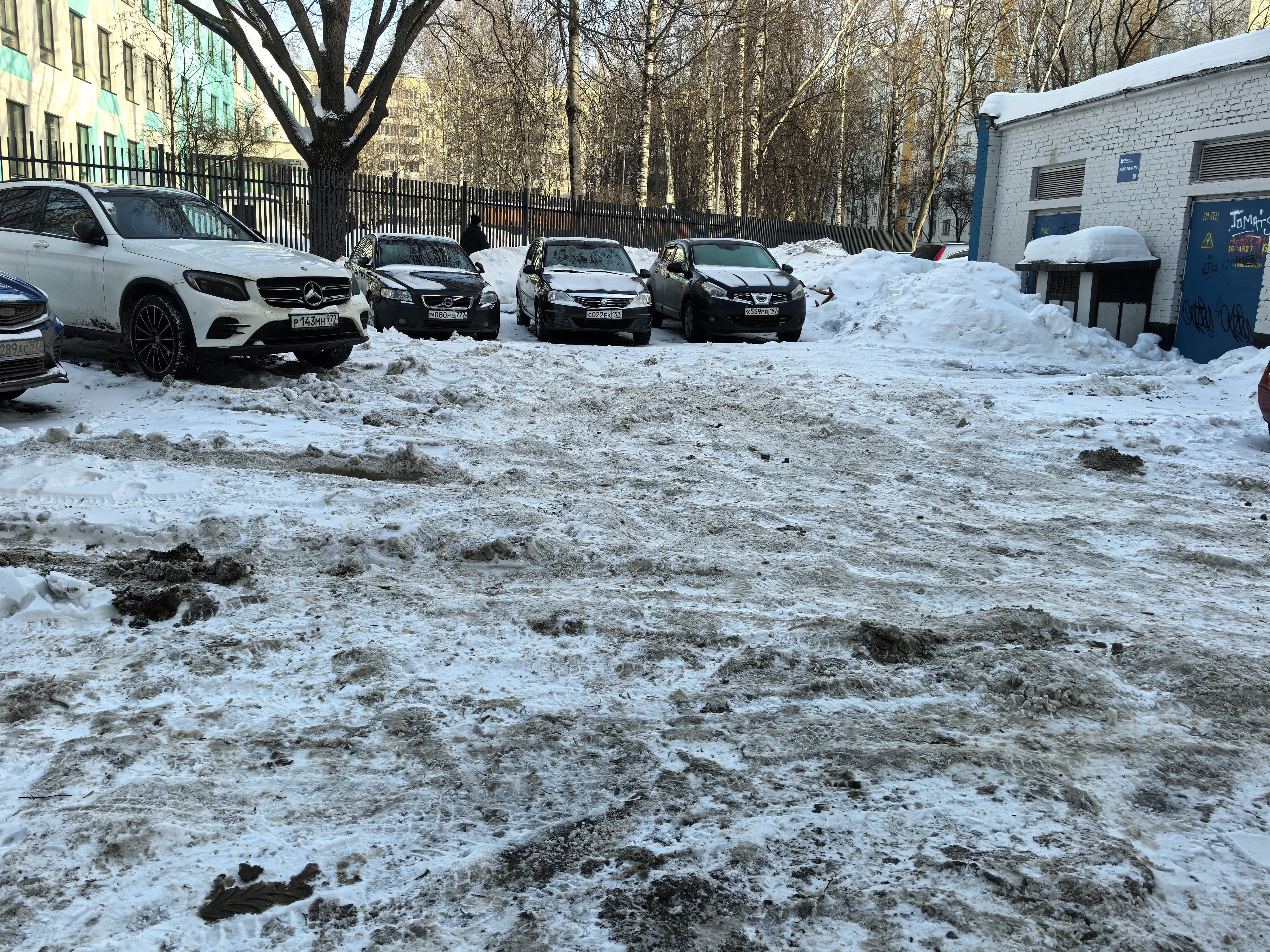 Дворовая территория с припаркованными автомобилями, покрытая снегом и грязью. Видны жилые дома и забор.