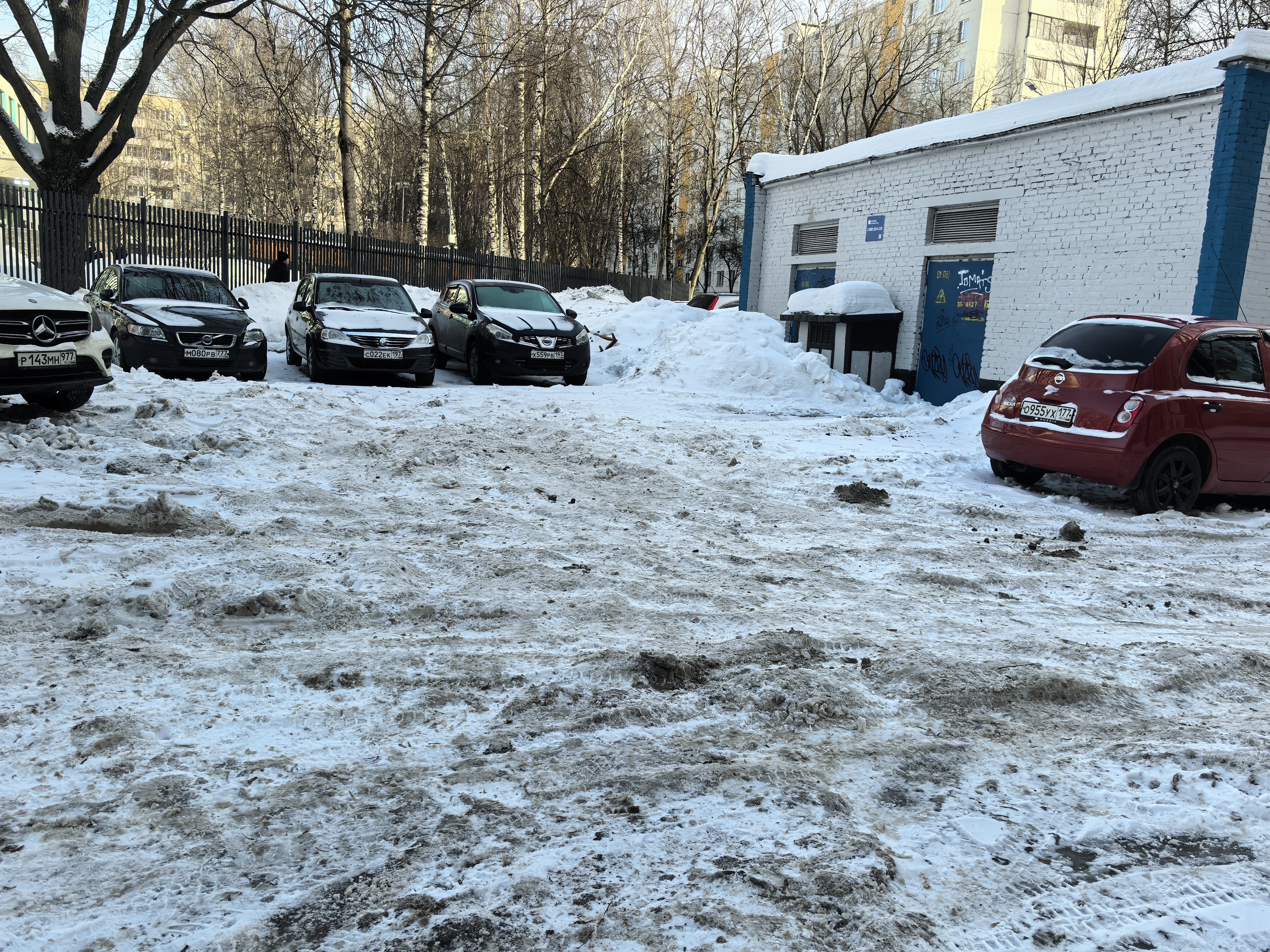 Дворовая территория с парковкой, покрытая снегом и грязью. Видны припаркованные автомобили, забор и гаражное строение. Дорога не имеет разметки, бордюров или пешеходных переходов.
