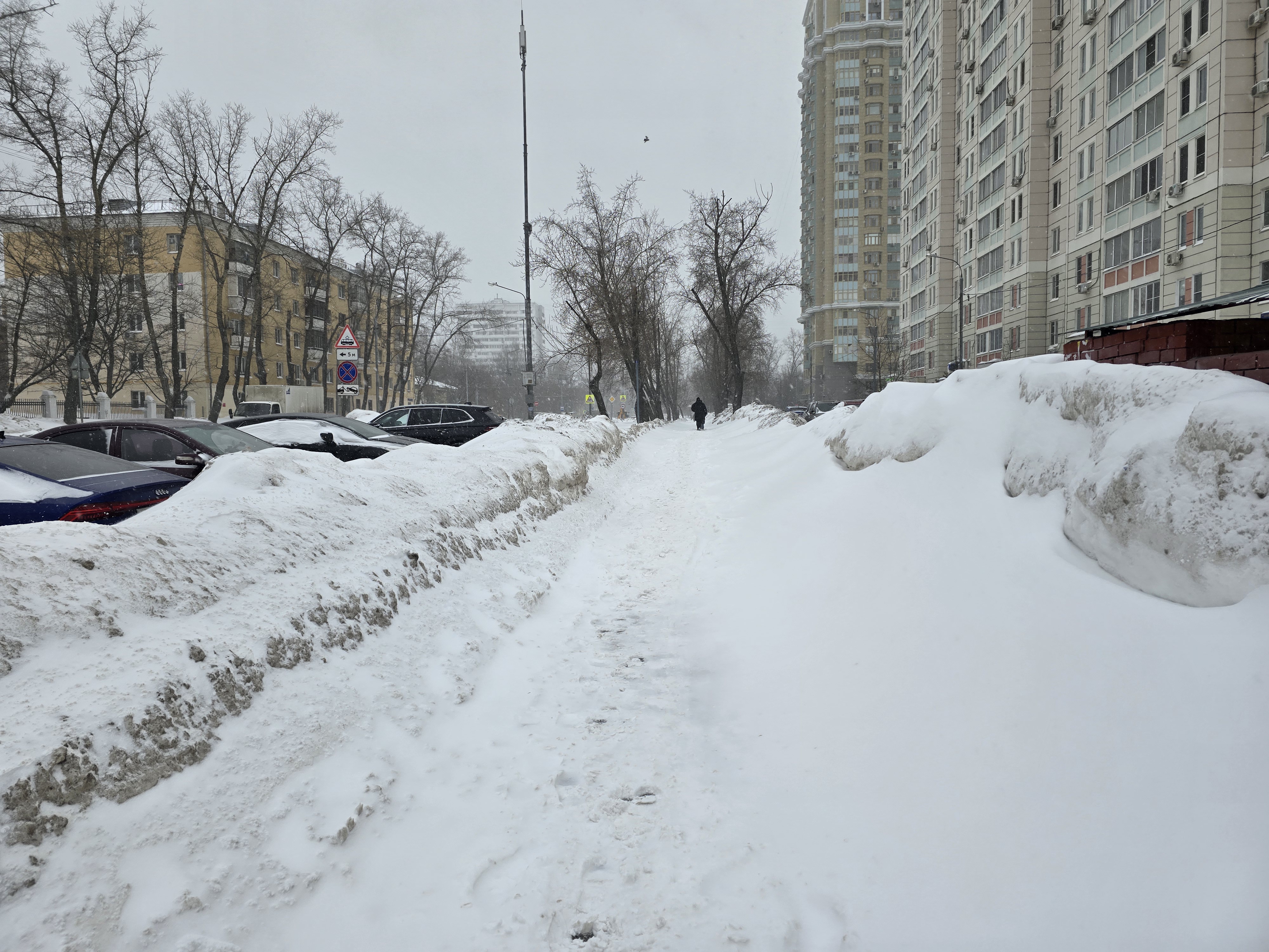 Участок дороги с пешеходным тротуаром, полностью покрытым снегом. Справа видны высокие жилые дома, слева — припаркованные автомобили и деревья. Пешеходного перехода на видимом участке нет.