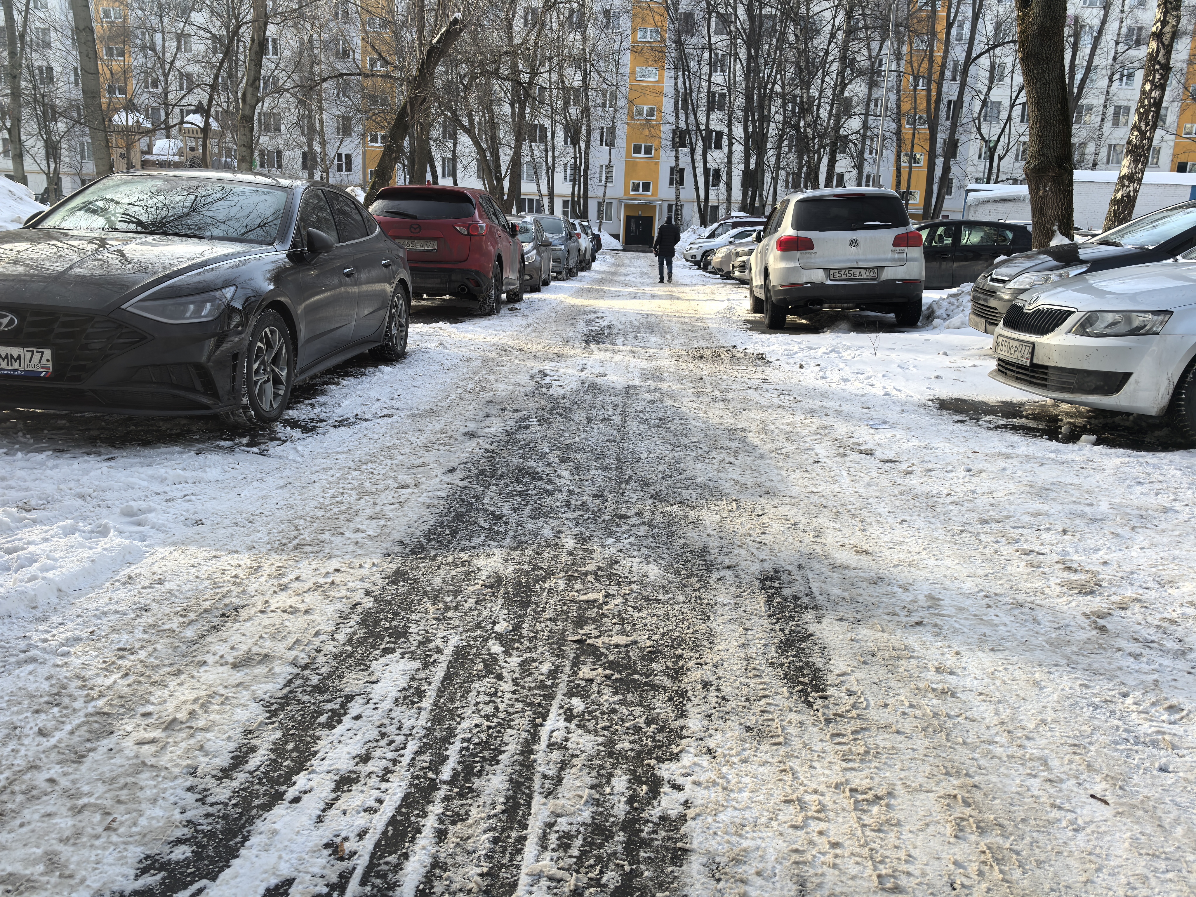 Дворовая территория с двусторонним движением, покрытая снегом и льдом. Парковка автомобилей вдоль дороги сужает проезжую часть, создавая узкий коридор для движения. На заднем плане виден жилой дом и пешеход, идущий по дороге.