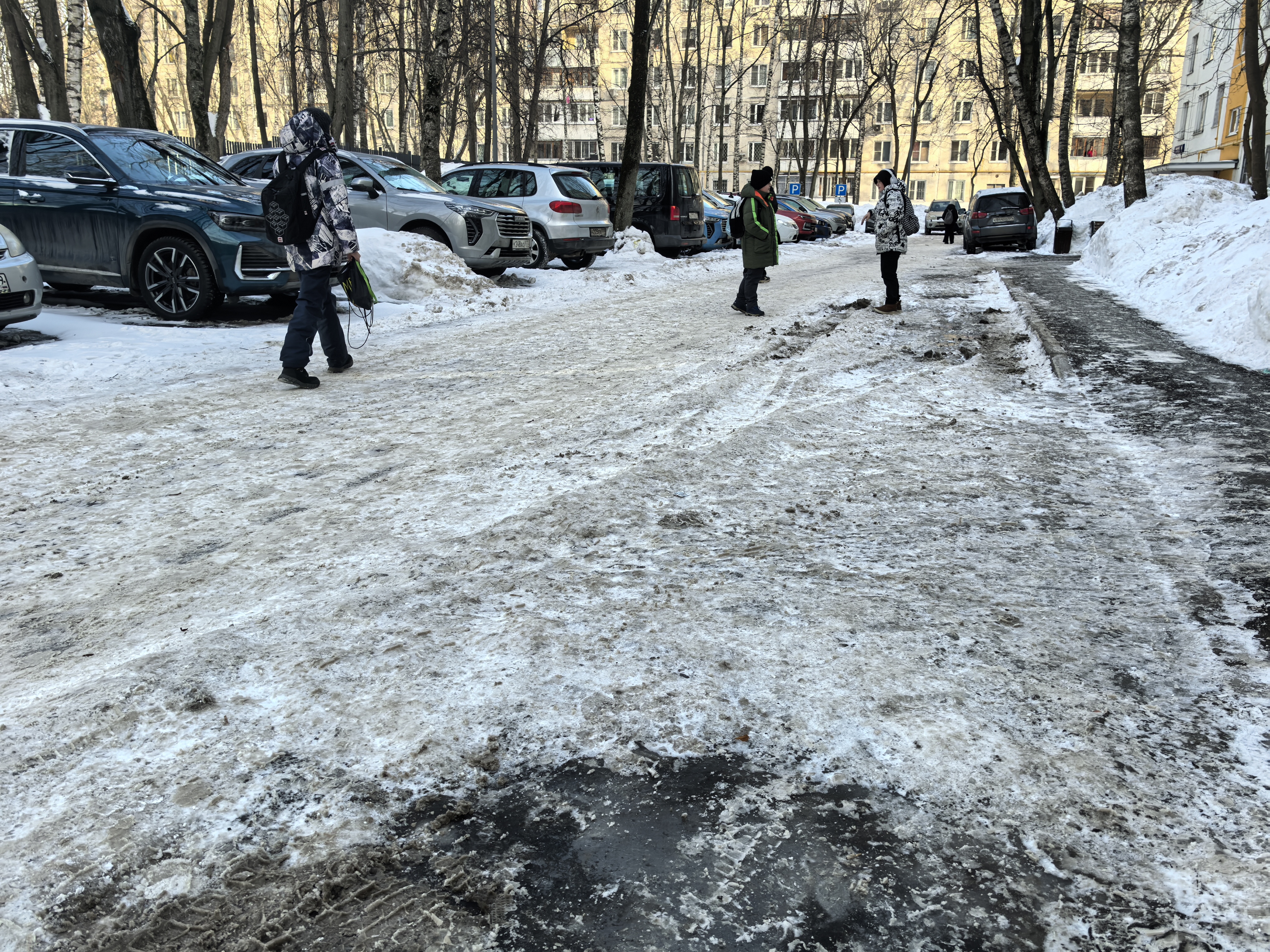 Дворовая территория с парковкой, покрытая снегом и льдом, с припаркованными автомобилями и пешеходами.