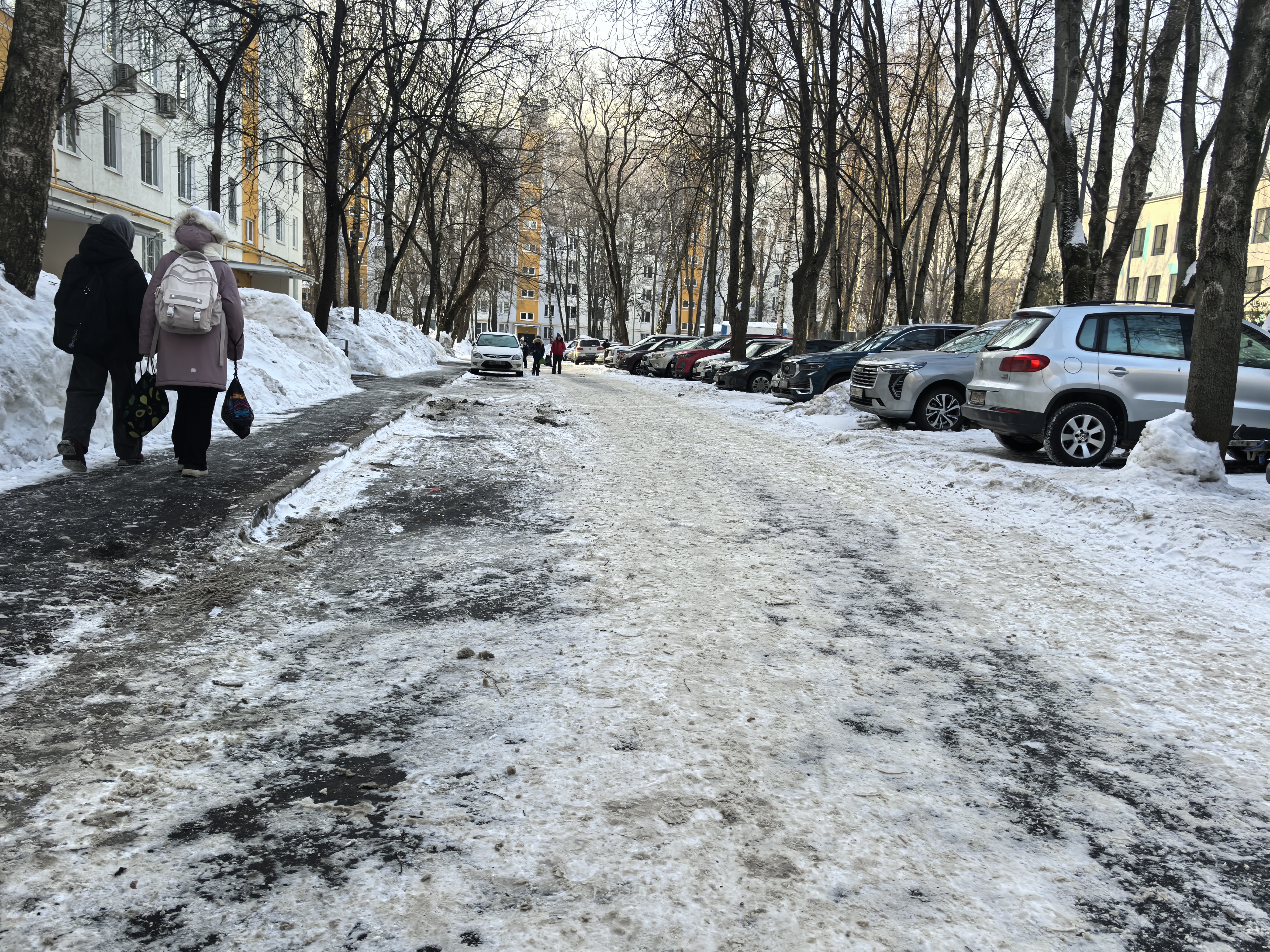 Дворовая территория с припаркованными автомобилями, покрытая снегом и льдом. Видны пешеходы на тротуаре и проезжая часть, где нет разметки.