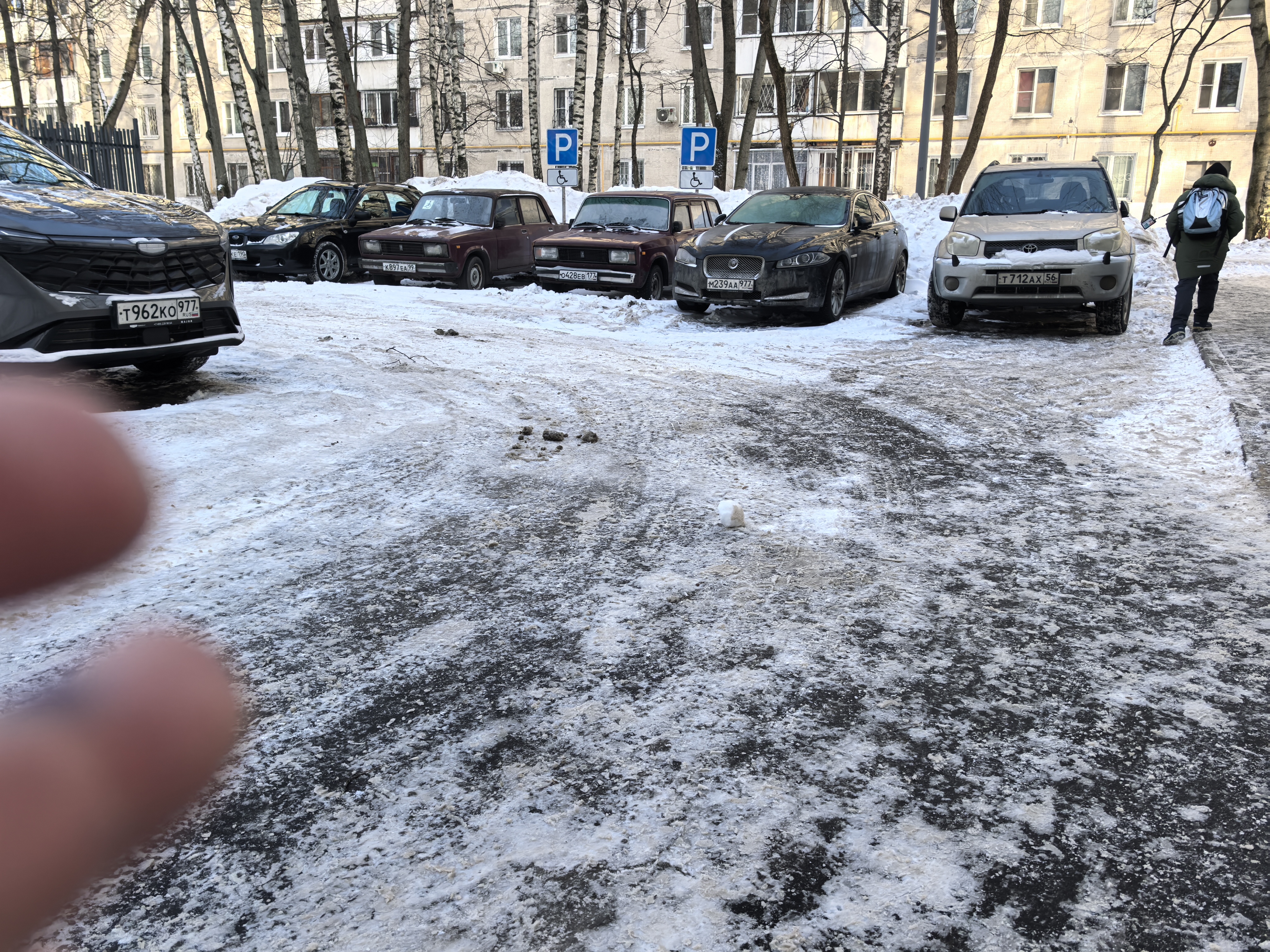 Дворовая территория с парковкой у жилых домов, покрытая снегом и льдом. Видны припаркованные автомобили и пешеход.