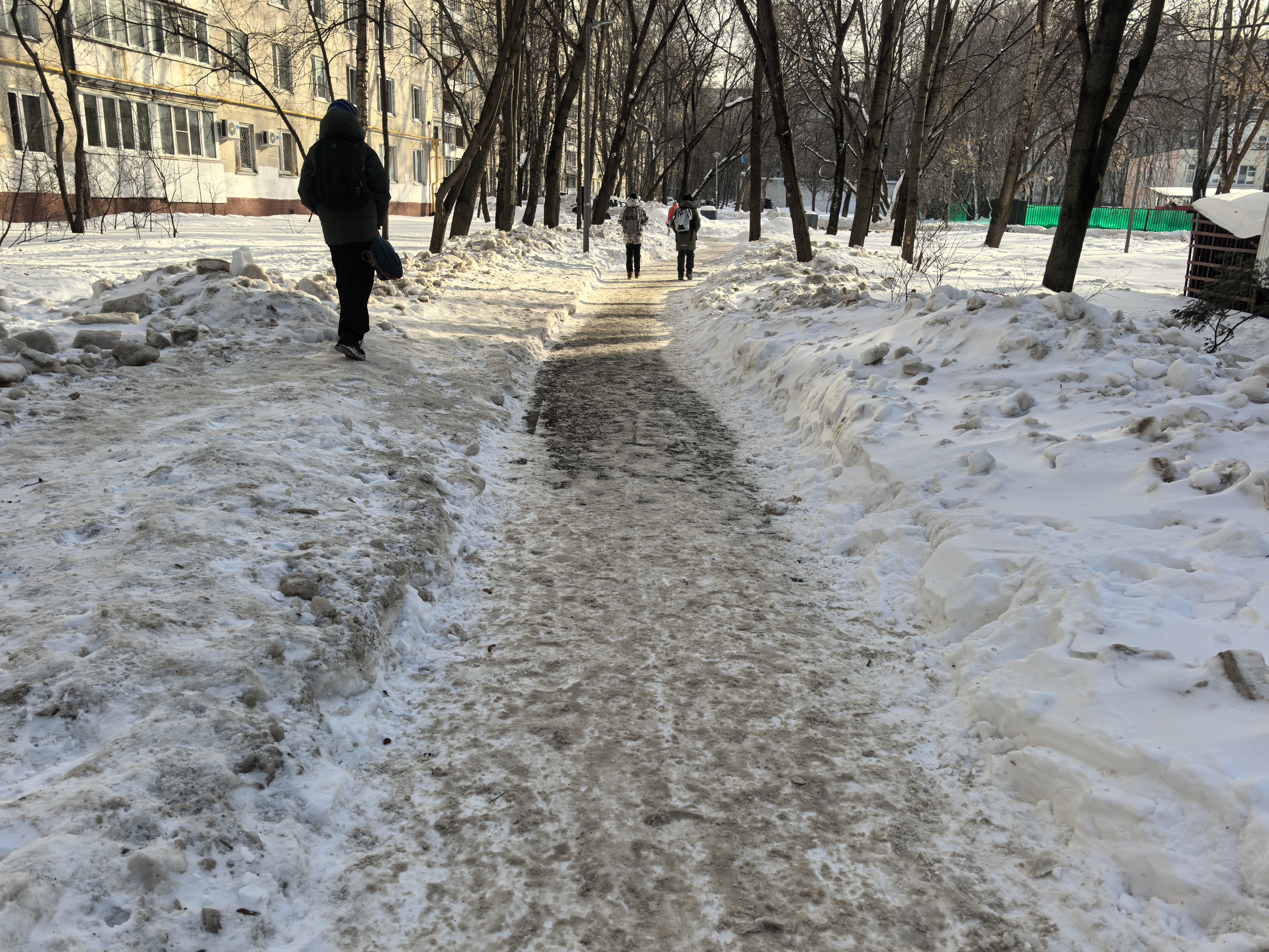 Дворовая территория или пешеходная дорожка в парке между жилыми домами, покрытая снегом и льдом. Видны люди, идущие по тропе.