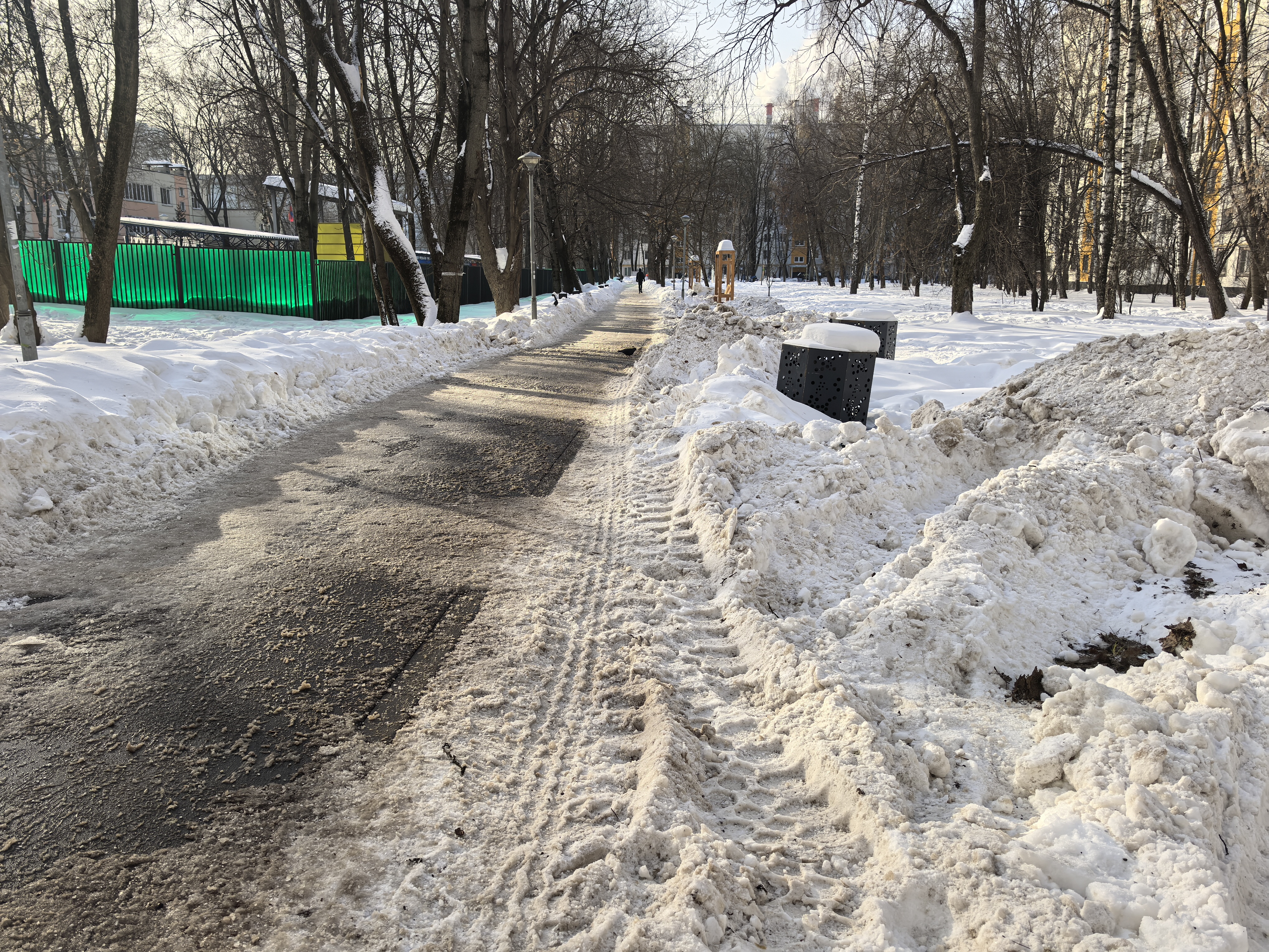 Дворовая территория или парк, зимний период. Дорога представляет собой пешеходную дорожку, очищенную от снега, с припаркованными мусорными контейнерами и деревьями по бокам.