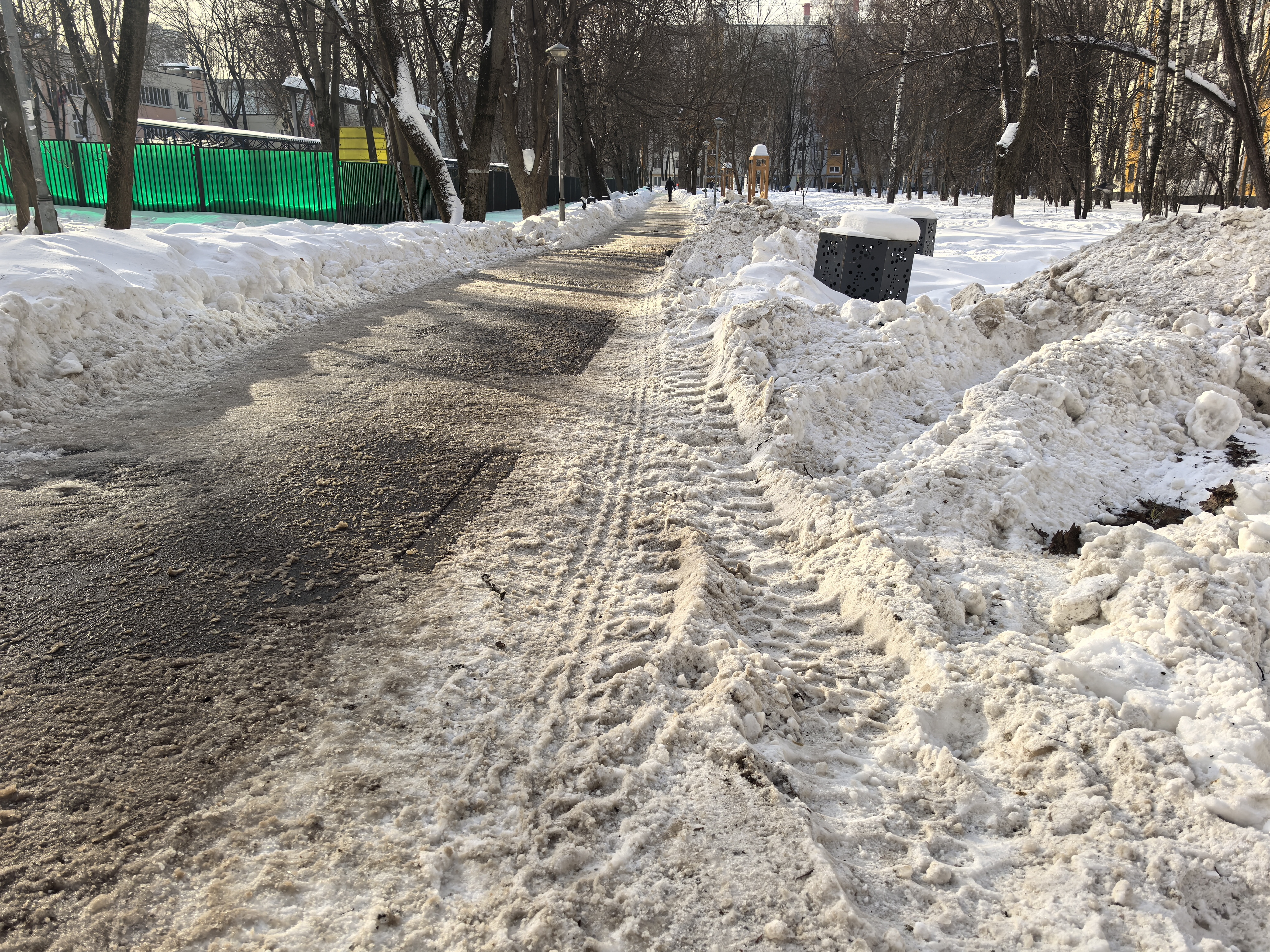 Дворовая территория или пешеходная дорожка в парке/сквере, очищенная от снега, с высокими сугробами по бокам. Присутствуют детские игровые элементы (деревянная башенка) и мусорные урны.