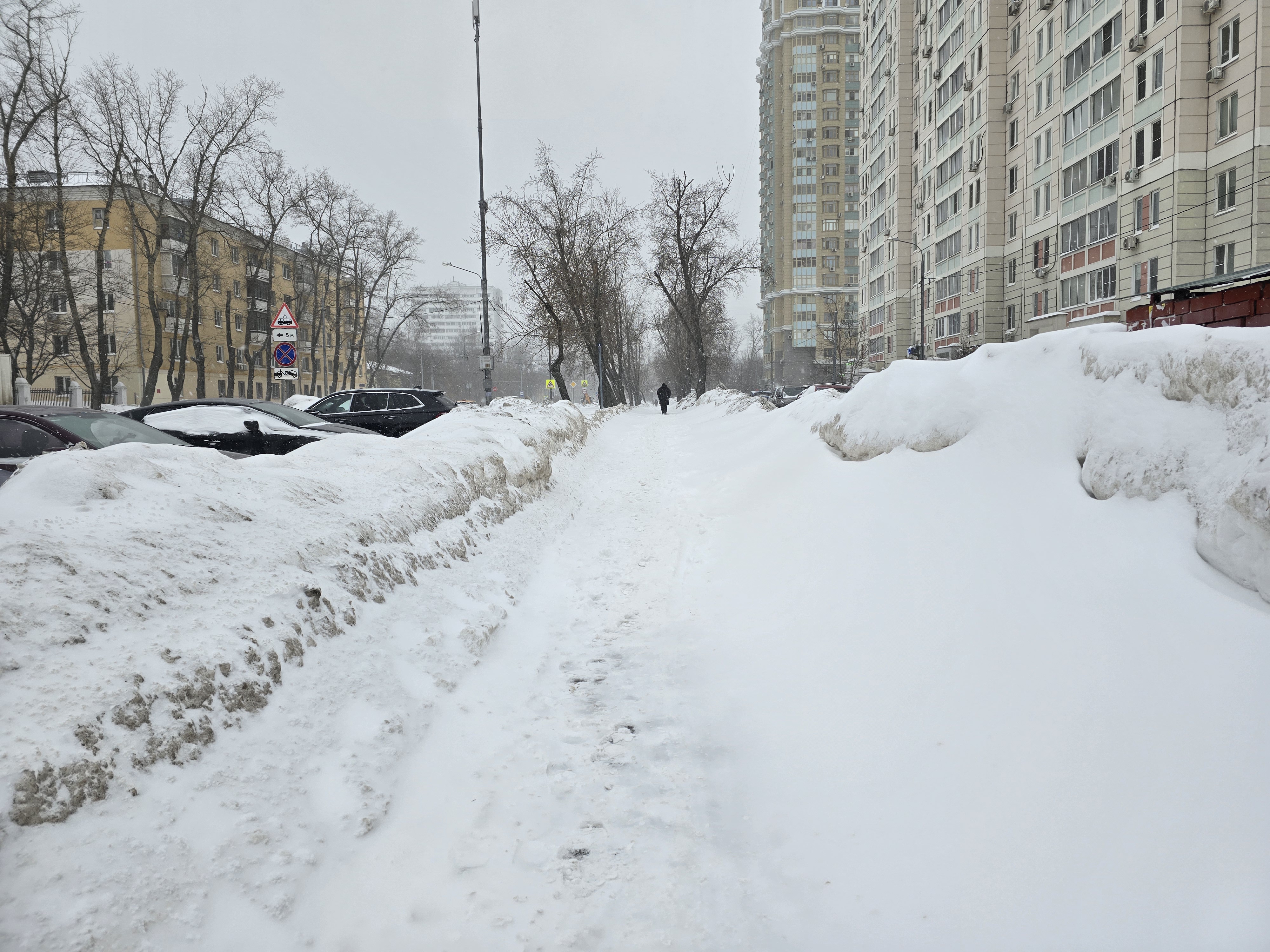 Участок городской улицы в зимний период. Тротуар полностью засыпан снегом, проезжая часть частично видна, припаркованы автомобили. Пешеходный переход отсутствует.