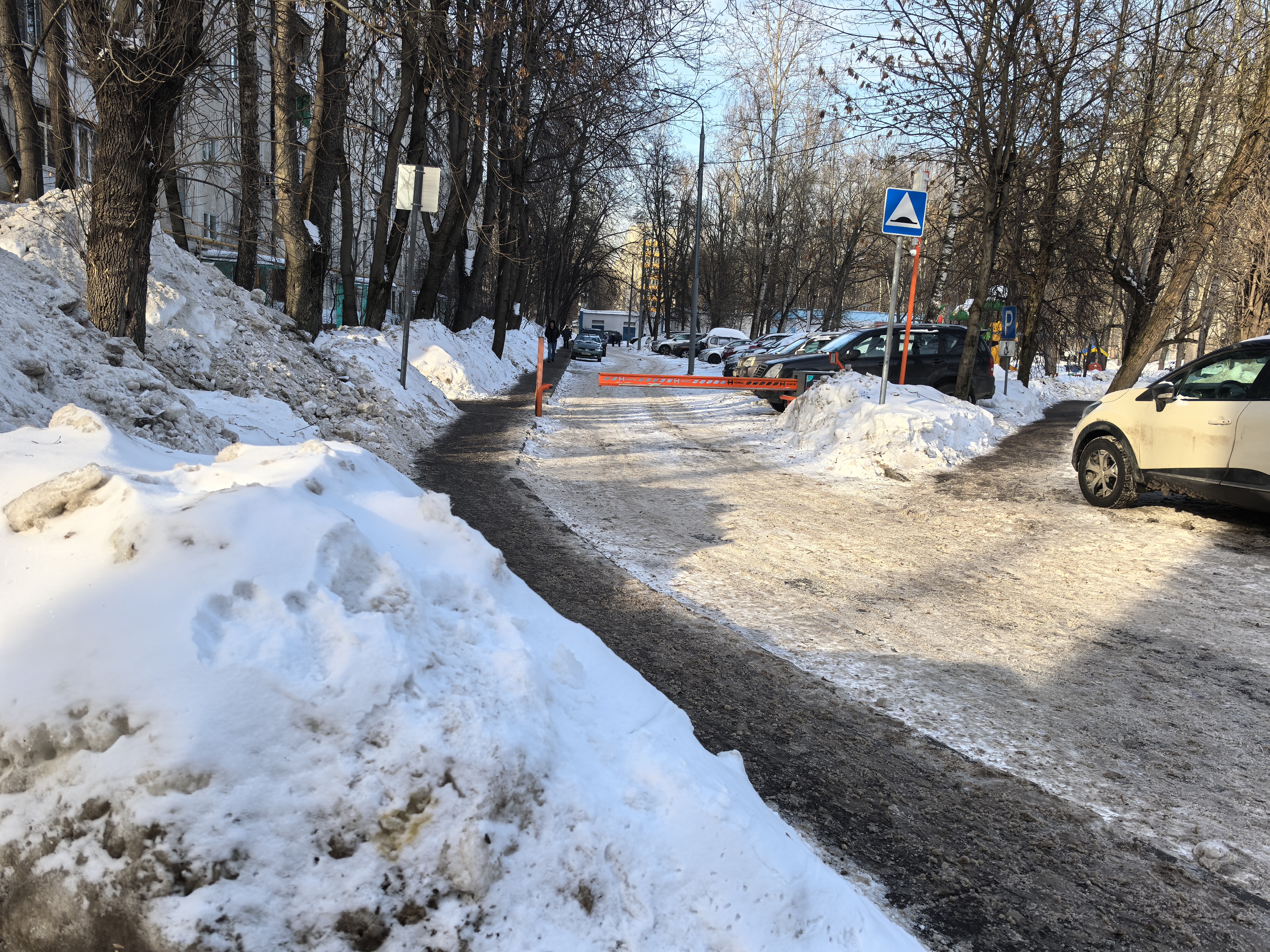 Дворовая территория с въездом на парковку, огороженная шлагбаумом. Дорога покрыта снегом и льдом, видны припаркованные автомобили.