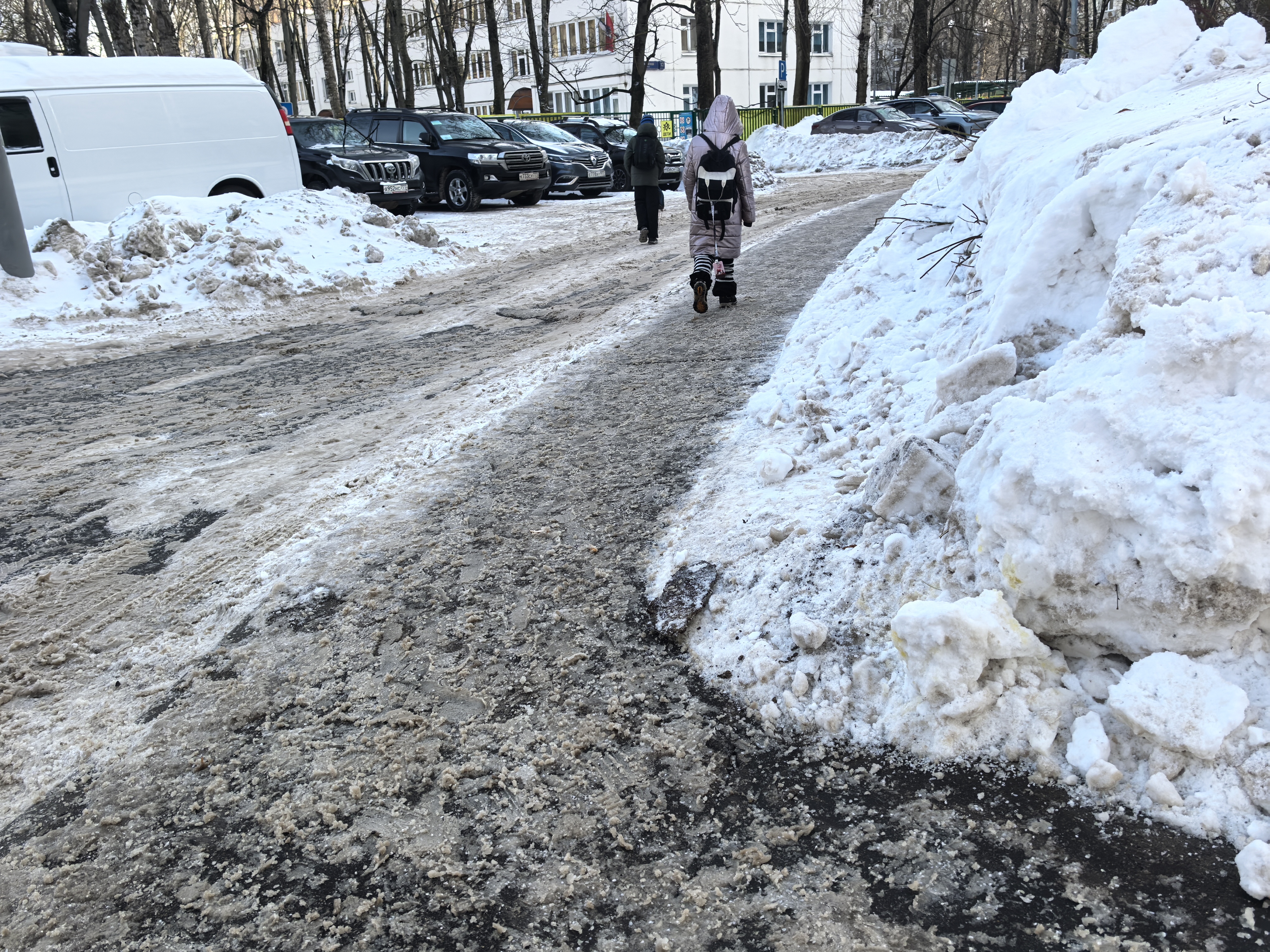 Дворовая территория с парковкой, покрытая снегом и ледяной кашей. Пешеходы идут по проезжей части, так как тротуары заблокированы сугробами.