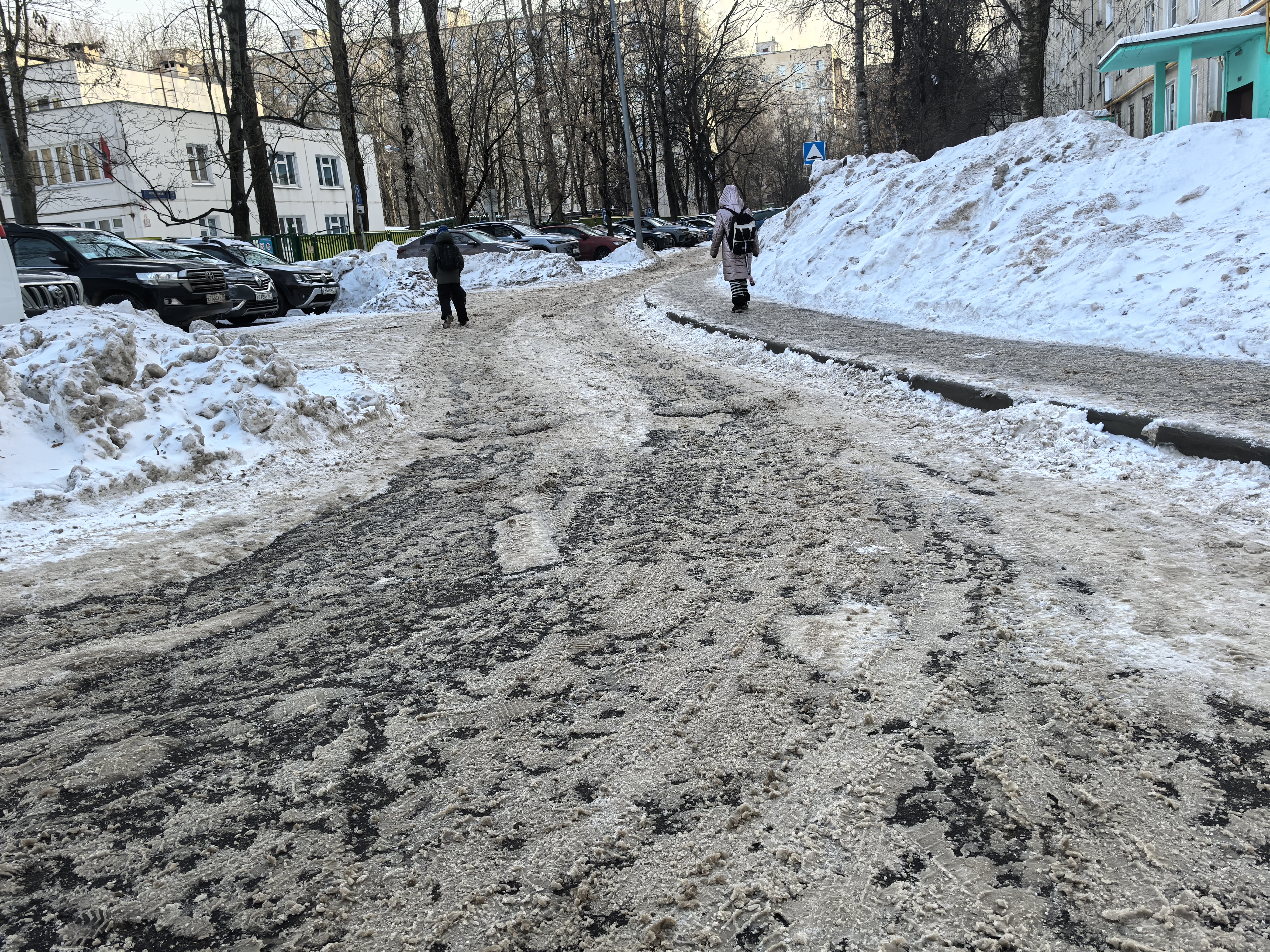 Участок дороги без пешеходного перехода, покрытый снегом и льдом. Тротуар справа очищен, но ограничен высокими сугробами.