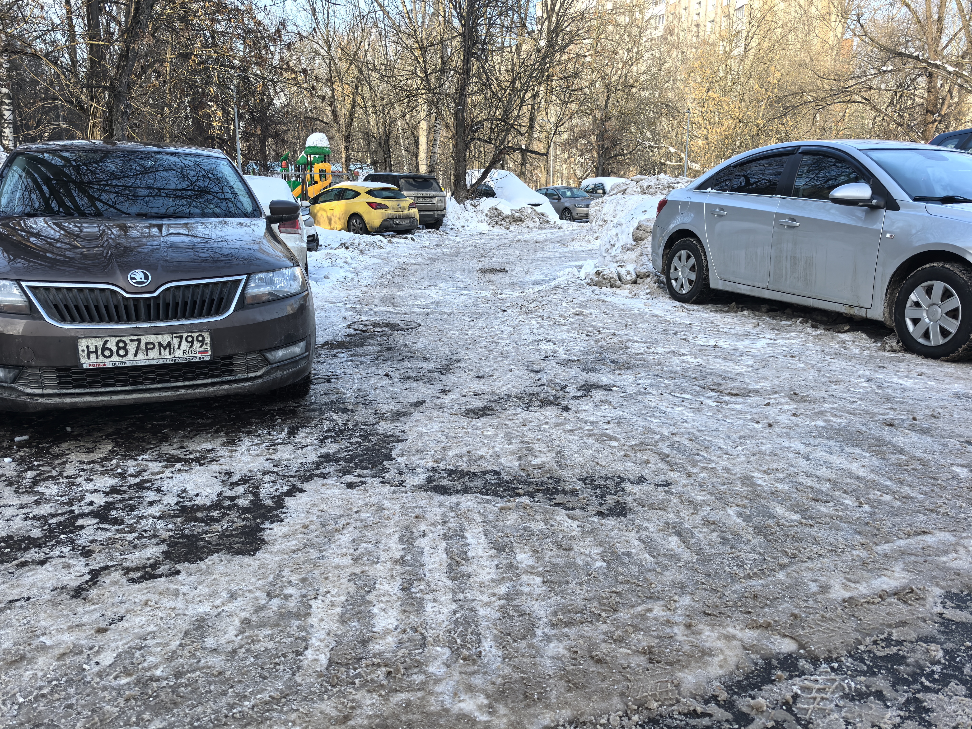 Дворовая территория с парковкой автомобилей, покрытая снегом и льдом. В глубине видна детская игровая площадка.