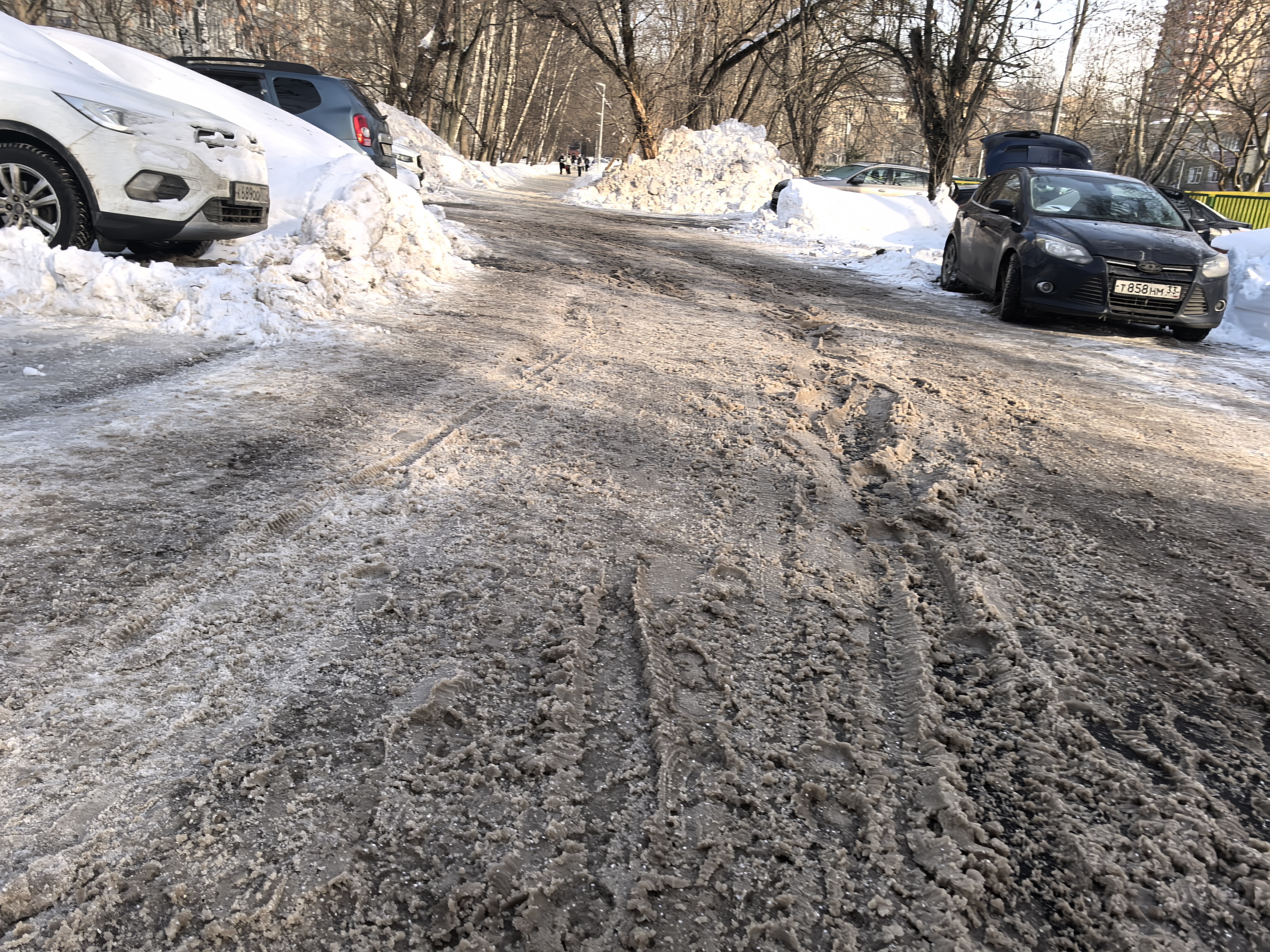 Участок проезжей части без обустроенного пешеходного перехода. Дорога покрыта снегом и льдом, припаркованные автомобили сужают проезжую часть и блокируют обзор.