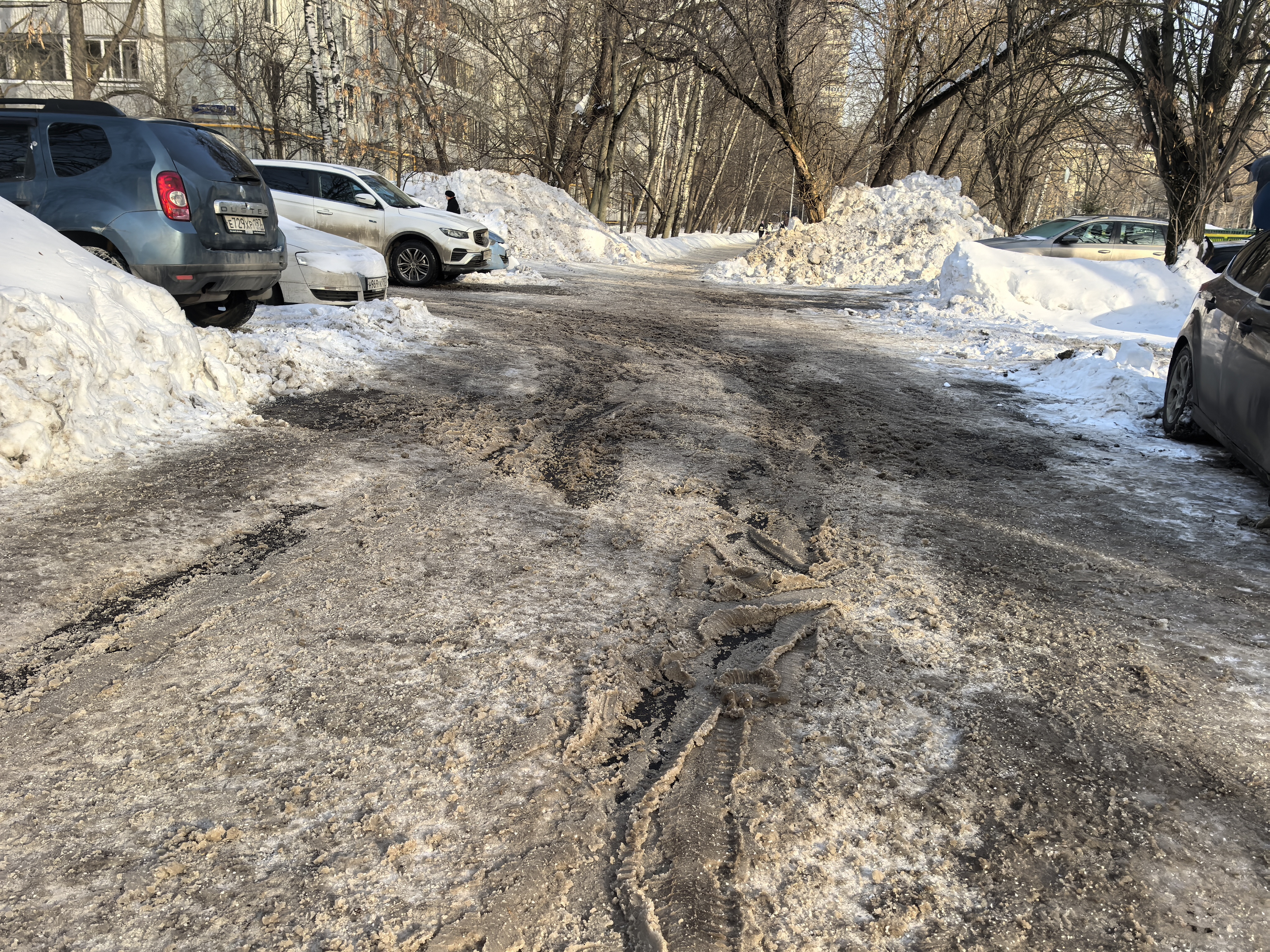 Дворовая территория, проезд между жилыми домами, покрытый снегом и ледяной кашей, с припаркованными автомобилями по бокам.