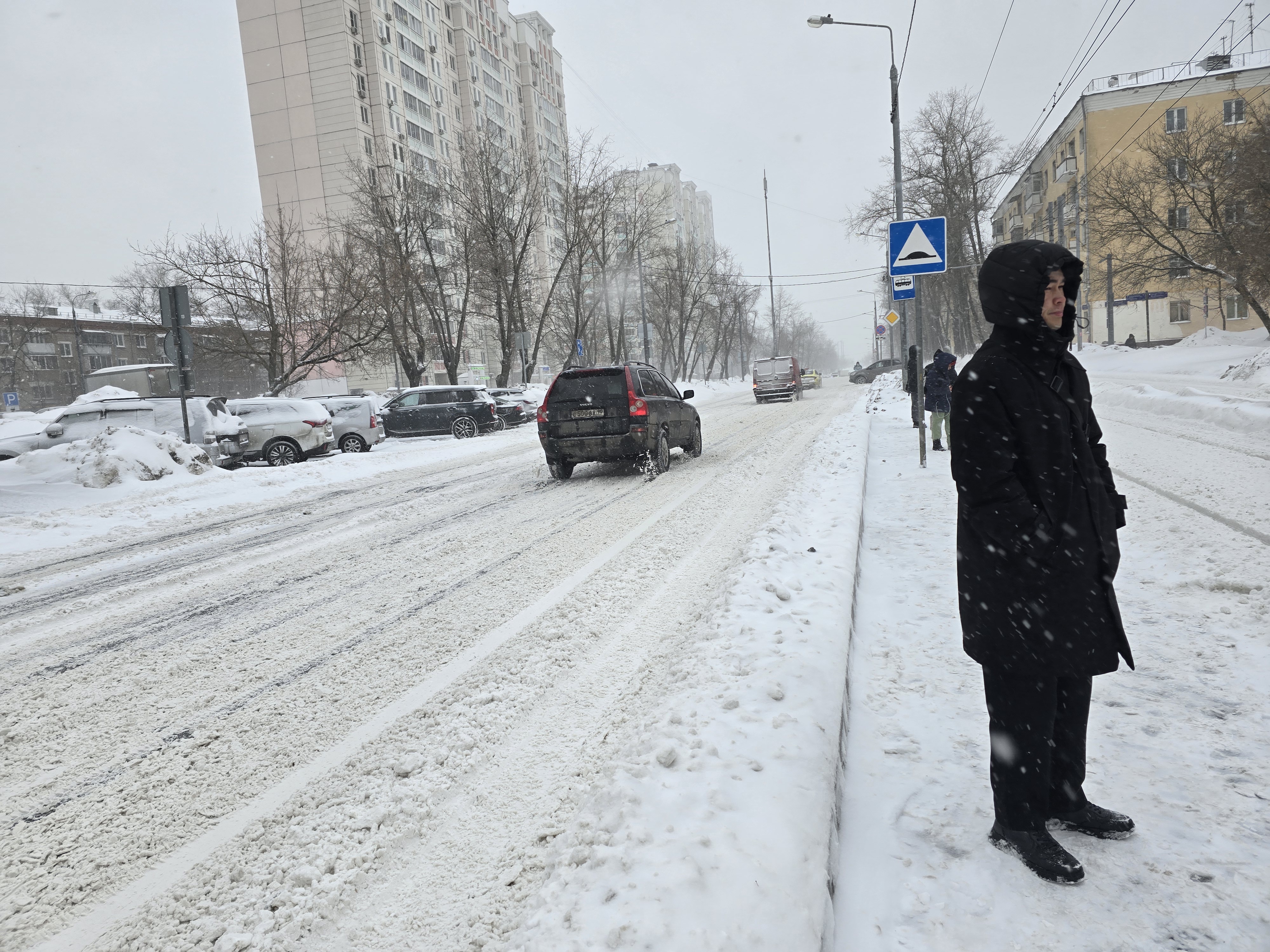 Участок городской дороги с тротуаром, покрытым снегом. Пешеходный переход отсутствует, виден знак «Дорожные неровности». На дороге движутся автомобили, припаркованные стоят на обочине.