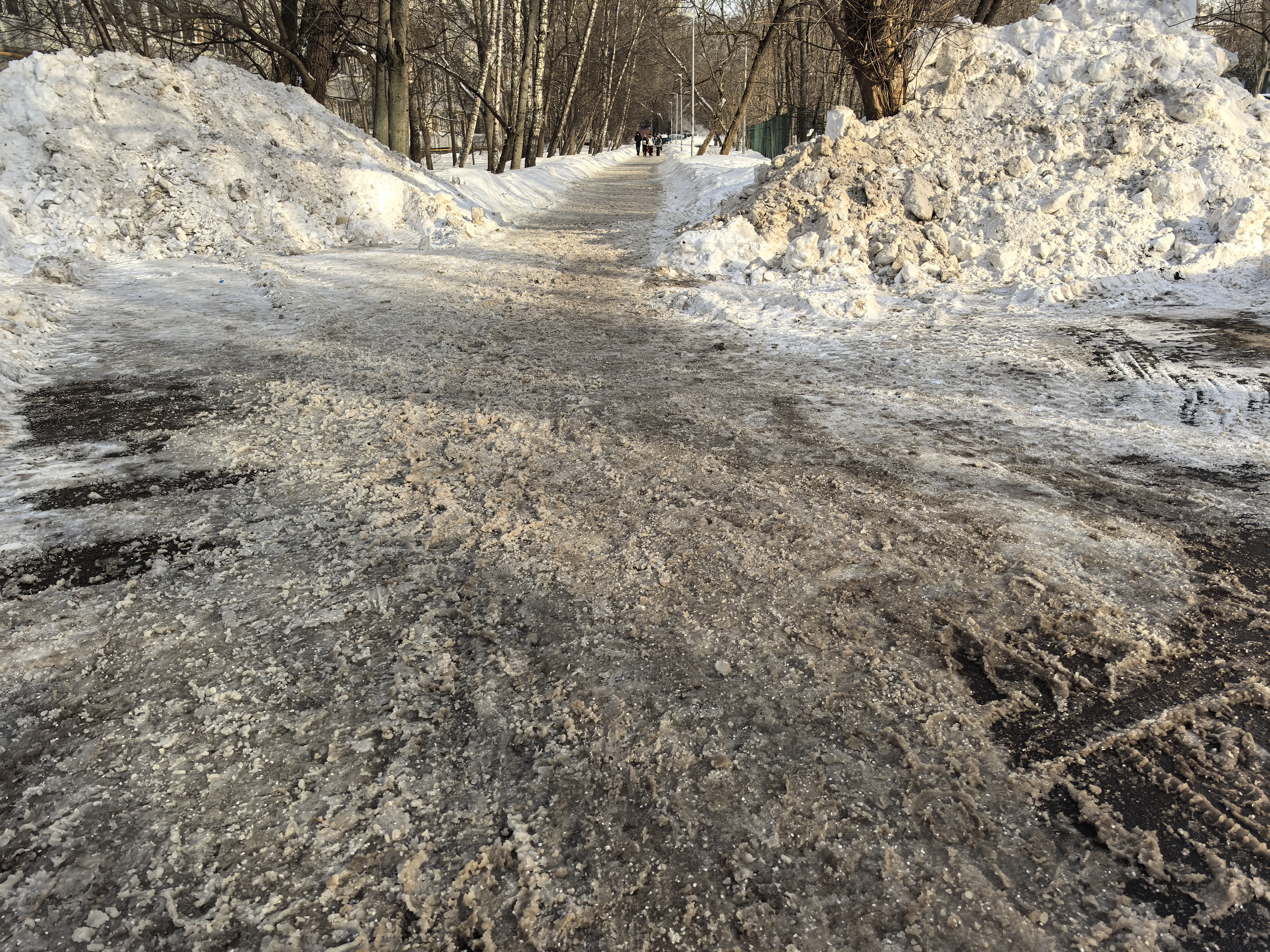 Дворовая территория или пешеходная дорожка в парке, покрытая снегом и льдом, с высокими сугробами по бокам.