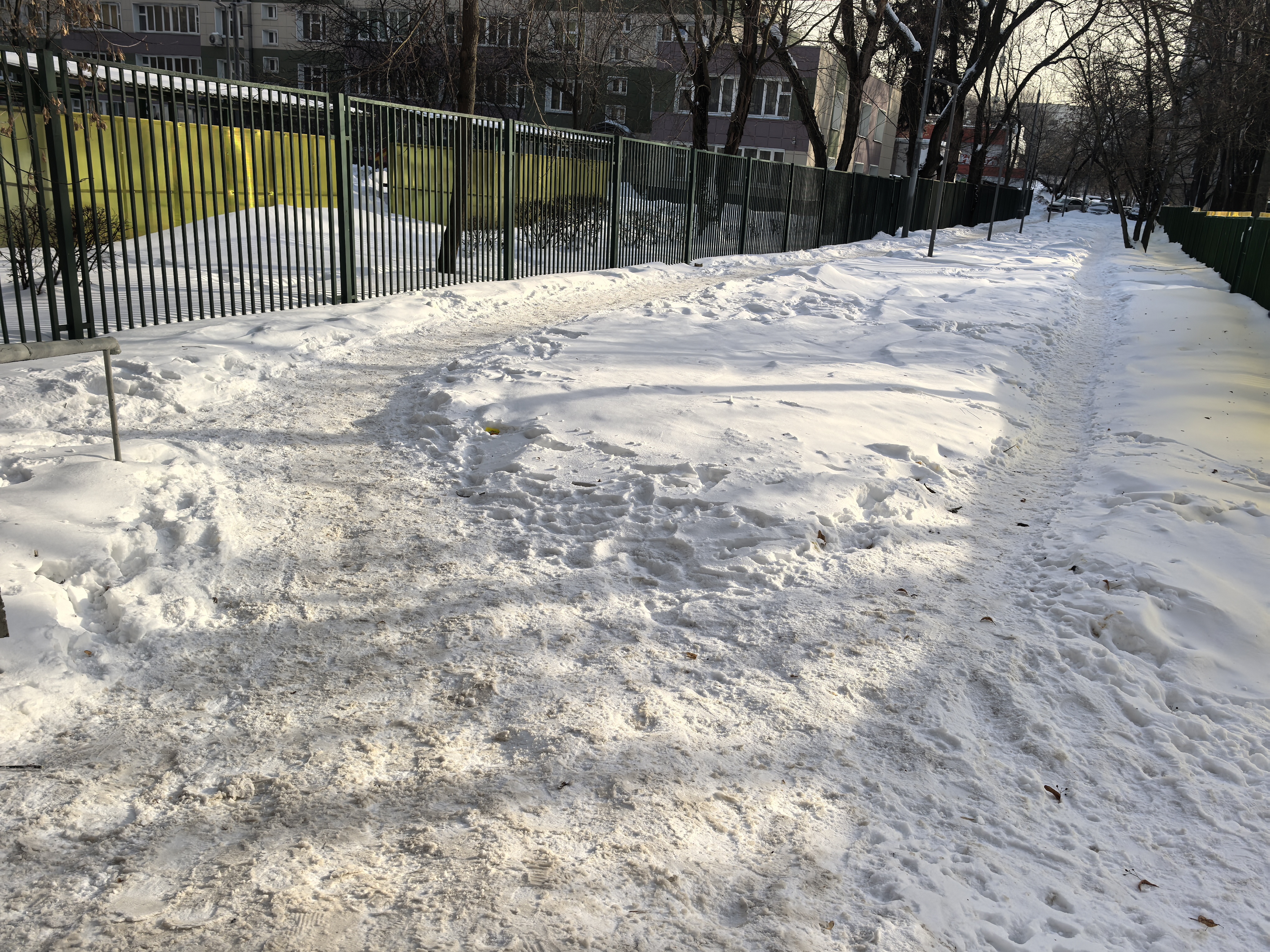 Участок пешеходного тротуара вдоль ограждения, полностью покрытый снегом. Проезжая часть не видна, но судя по расположению, это придорожная зона.