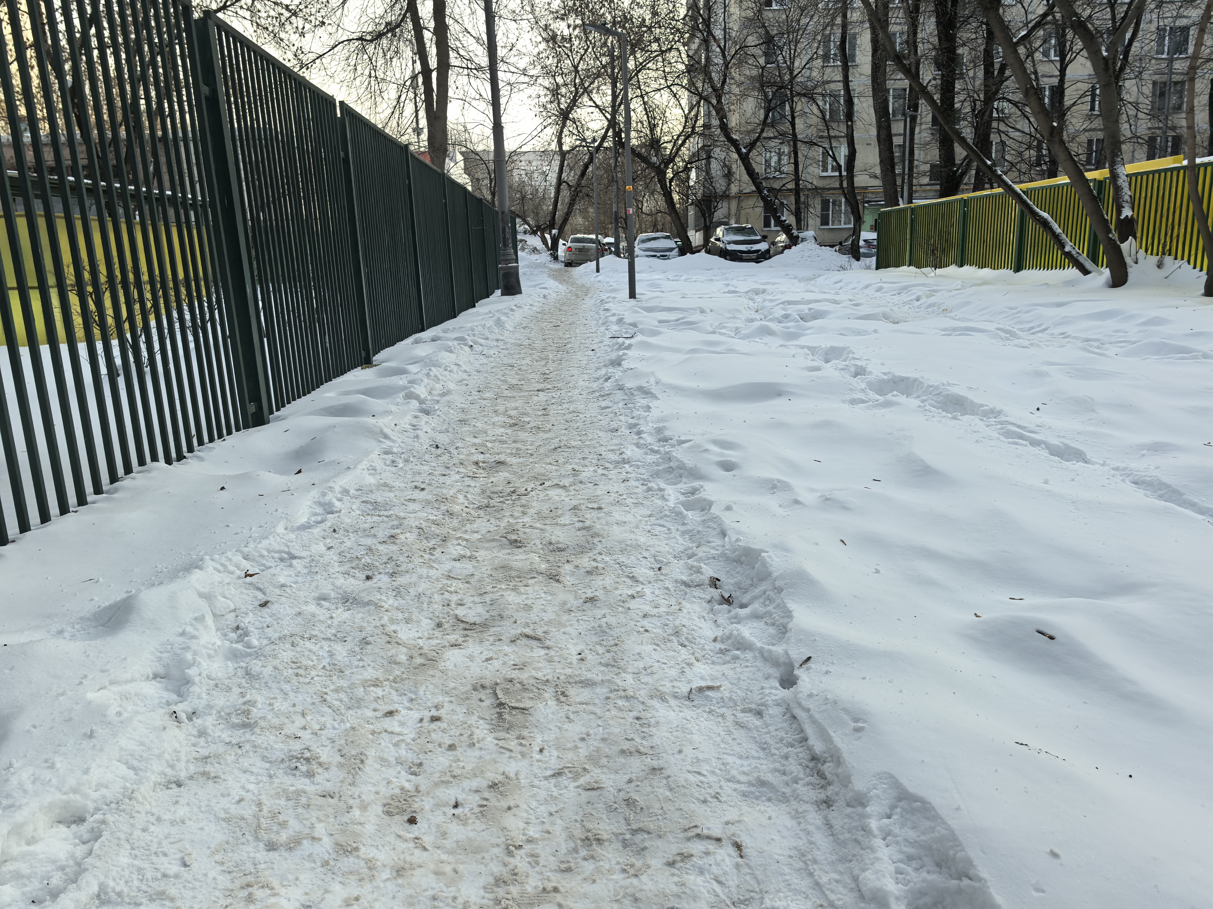 Зимний двор или территория между жилыми домами. Видна заснеженная дорожка (вероятно, тротуар или пешеходная зона), окруженная забором и деревьями. На заднем плане видны припаркованные автомобили, что характерно для дворовых территорий.
