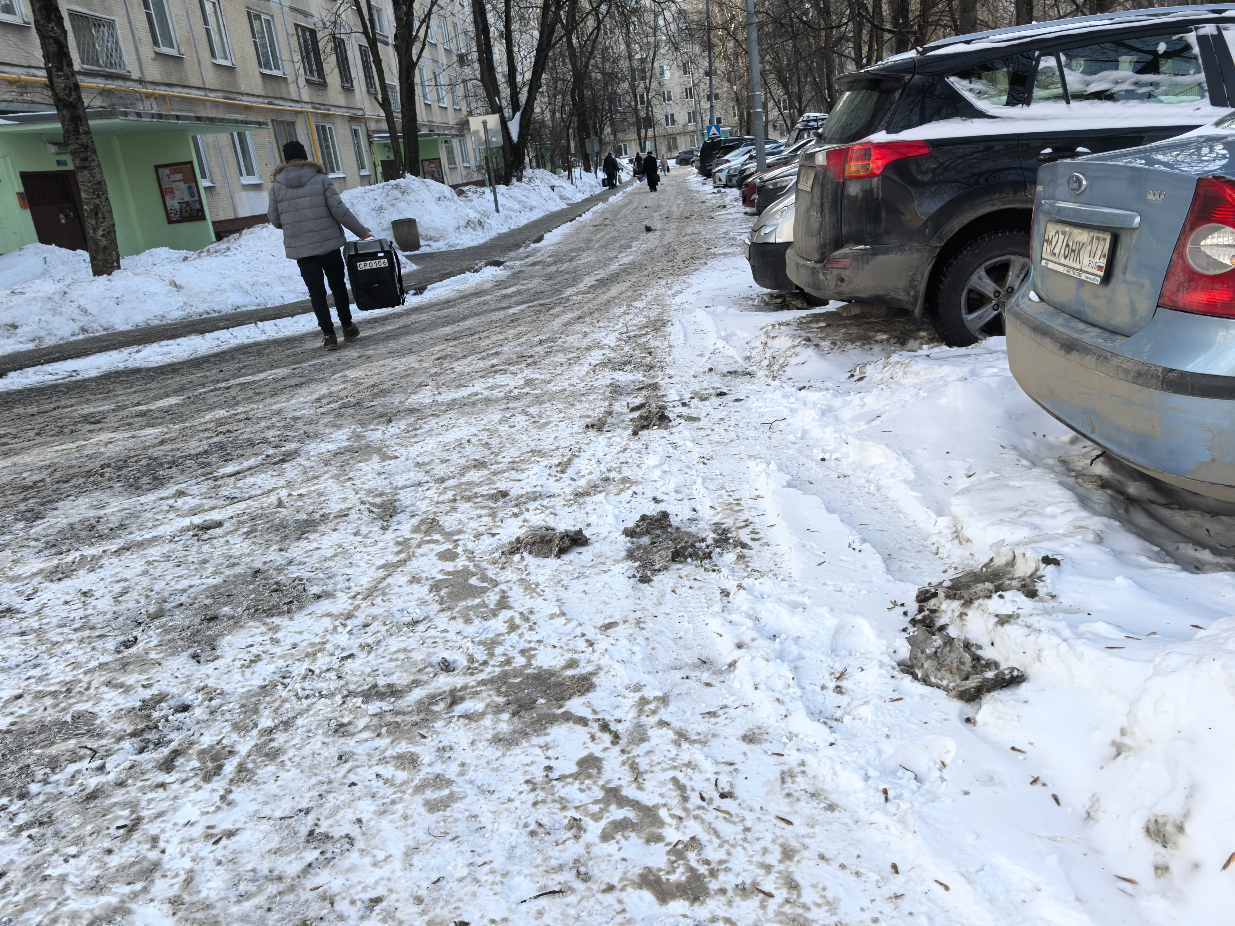 Участок дороги во дворе или прилегающей территории, покрытый снегом и грязью. Пешеходный переход отсутствует, тротуары частично засыпаны снегом, припаркованные автомобили стоят вдоль дороги.