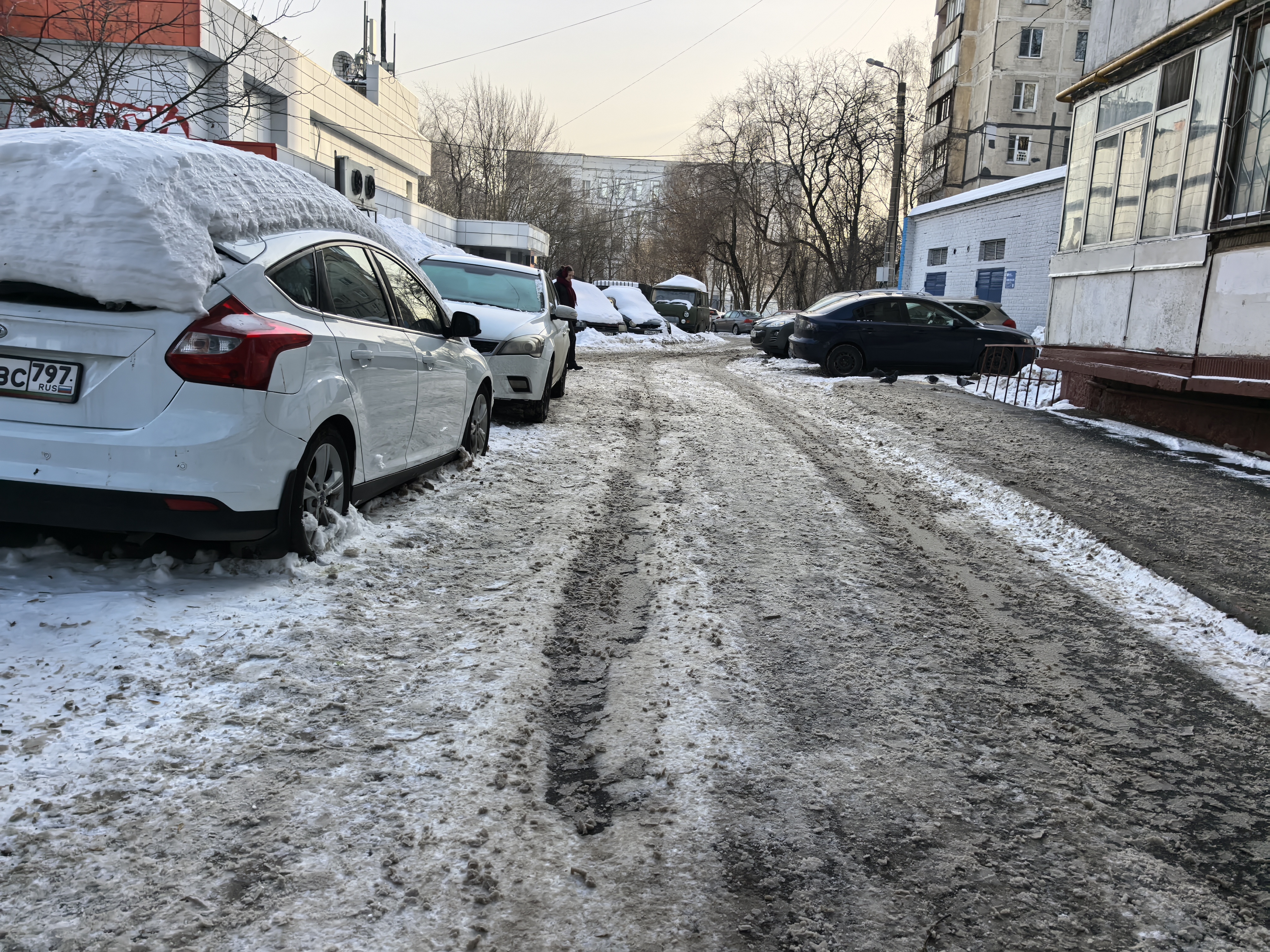 Дворовая территория с припаркованными автомобилями, покрытая снегом и грязью. Проезжая часть узкая, без разметки и пешеходных переходов. Видны жилые дома и деревья.