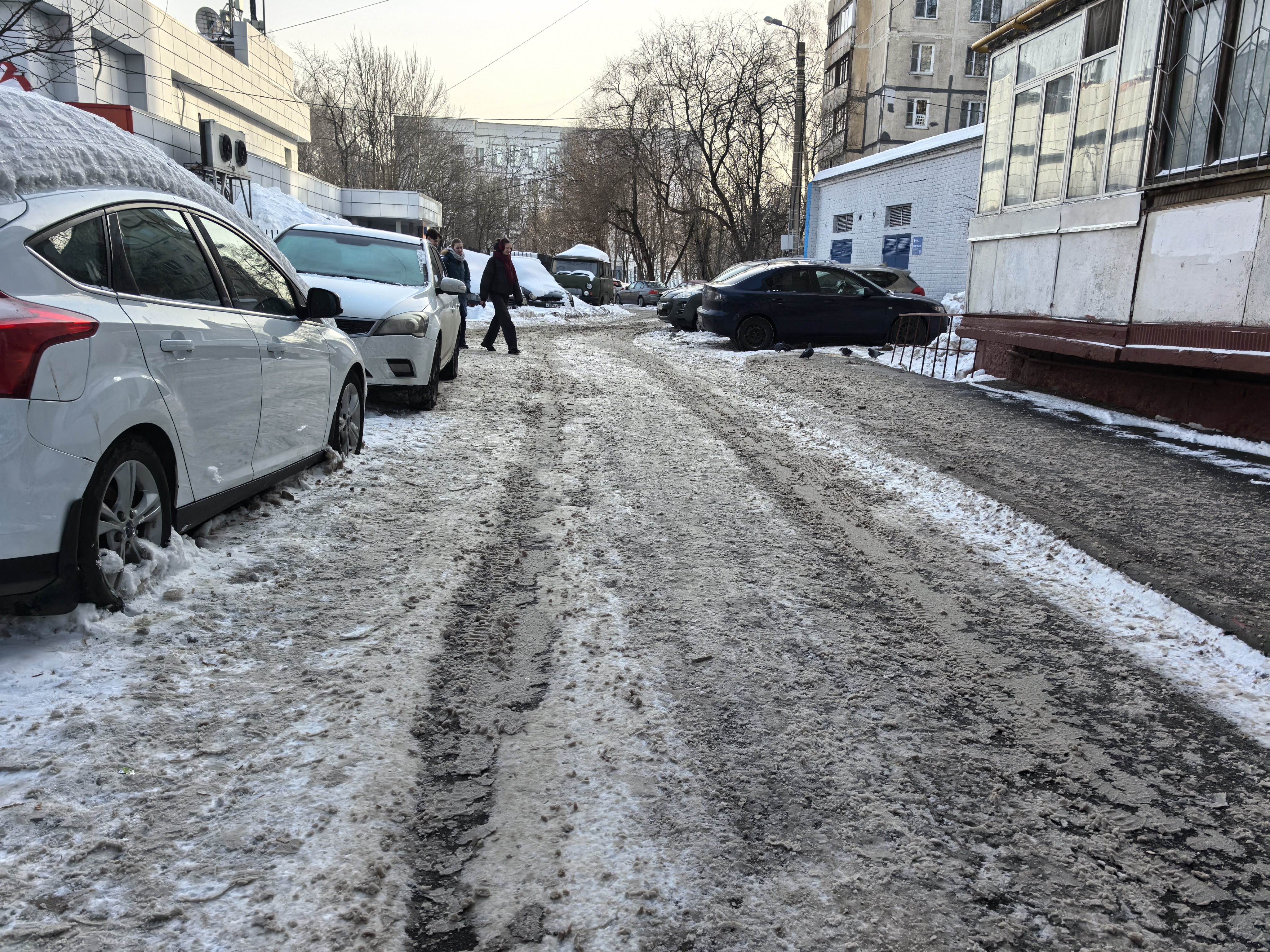 Дворовая территория с припаркованными автомобилями и пешеходами, проезжая часть покрыта снегом и грязью, тротуаров нет.