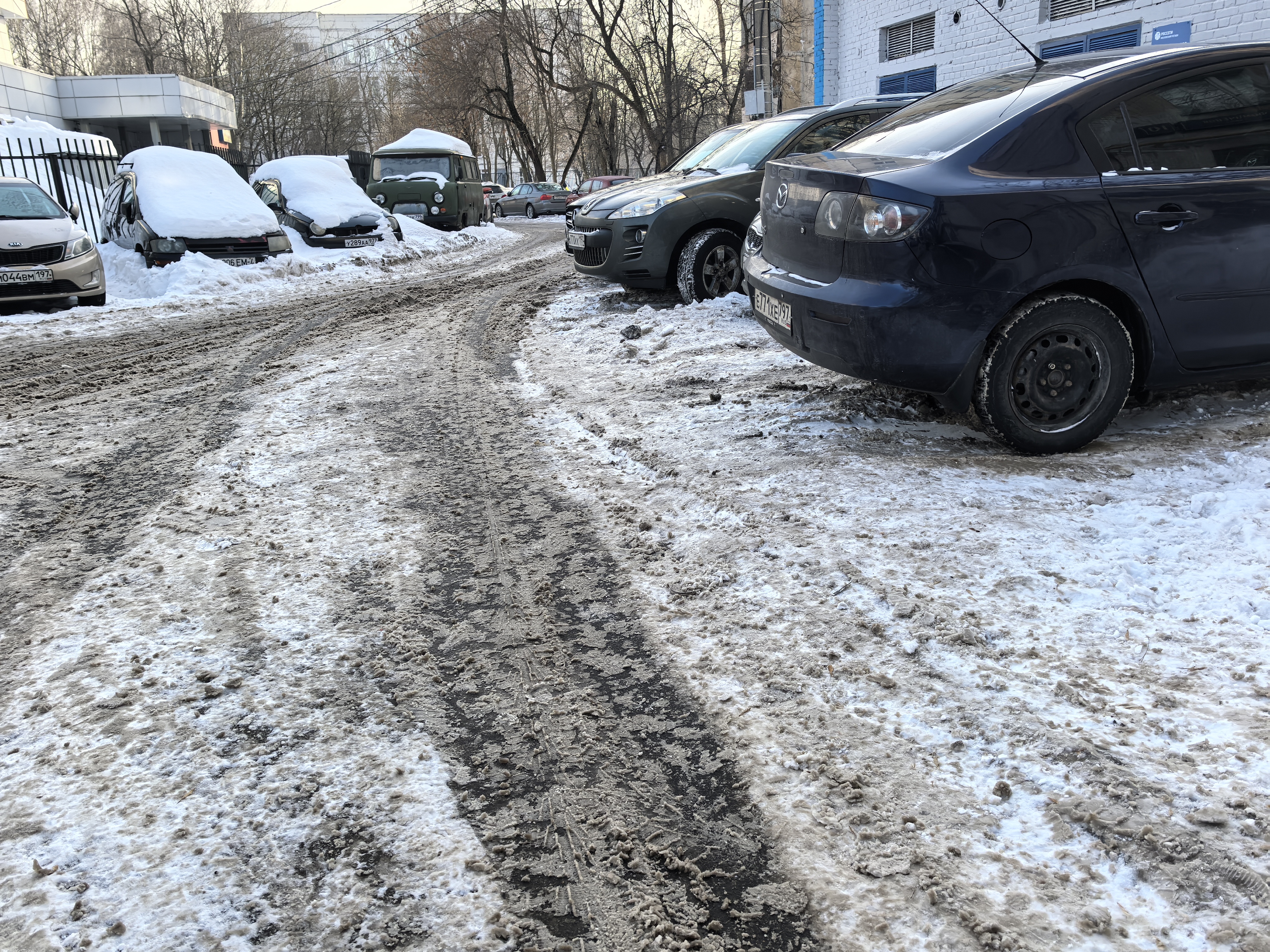 Дворовая территория с припаркованными автомобилями, проезжая часть покрыта снегом и грязью, тротуаров нет.