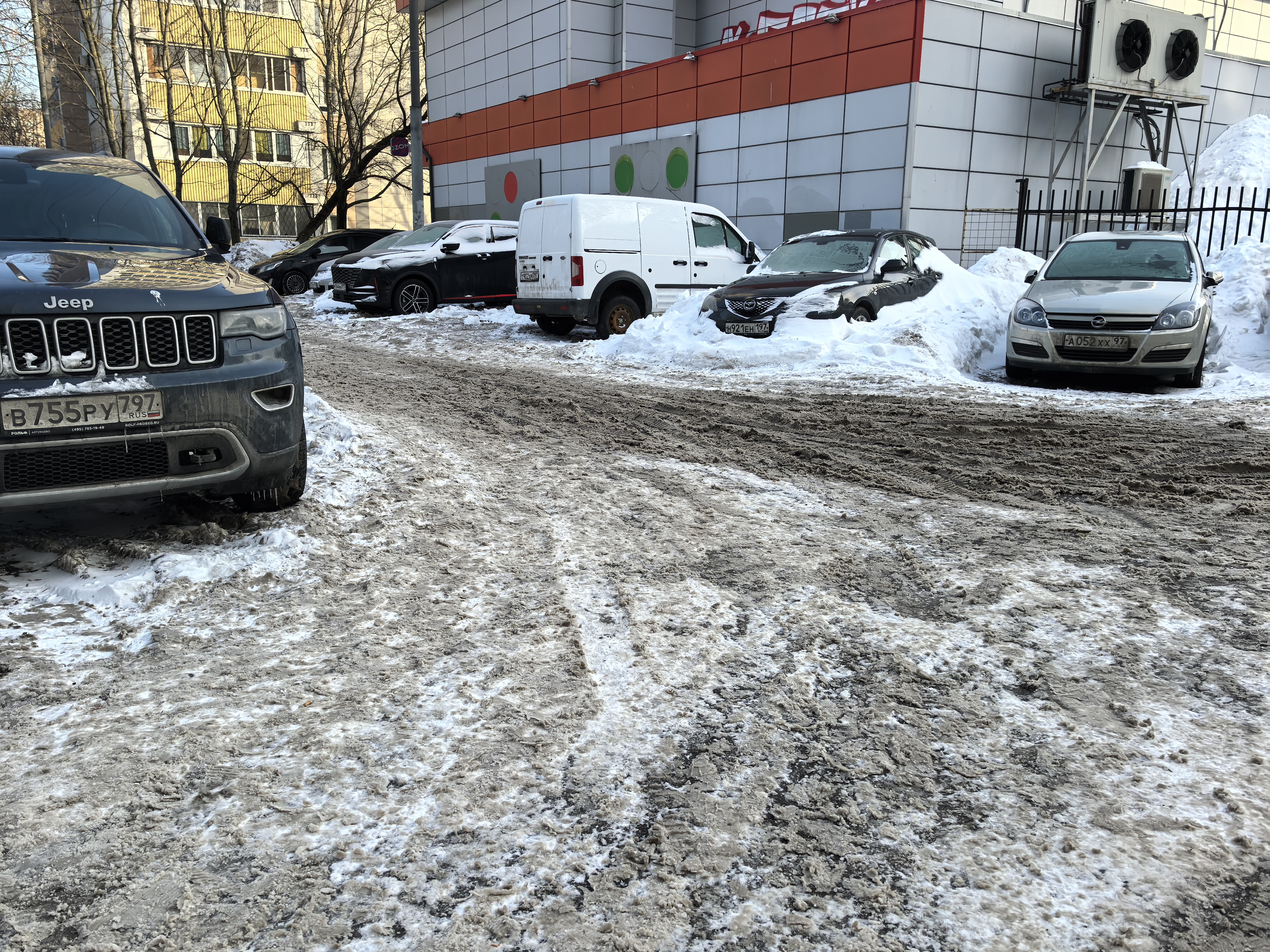 Дворовая территория или парковка у здания, покрытая снегом и грязью. Видны припаркованные автомобили и отсутствие тротуаров.