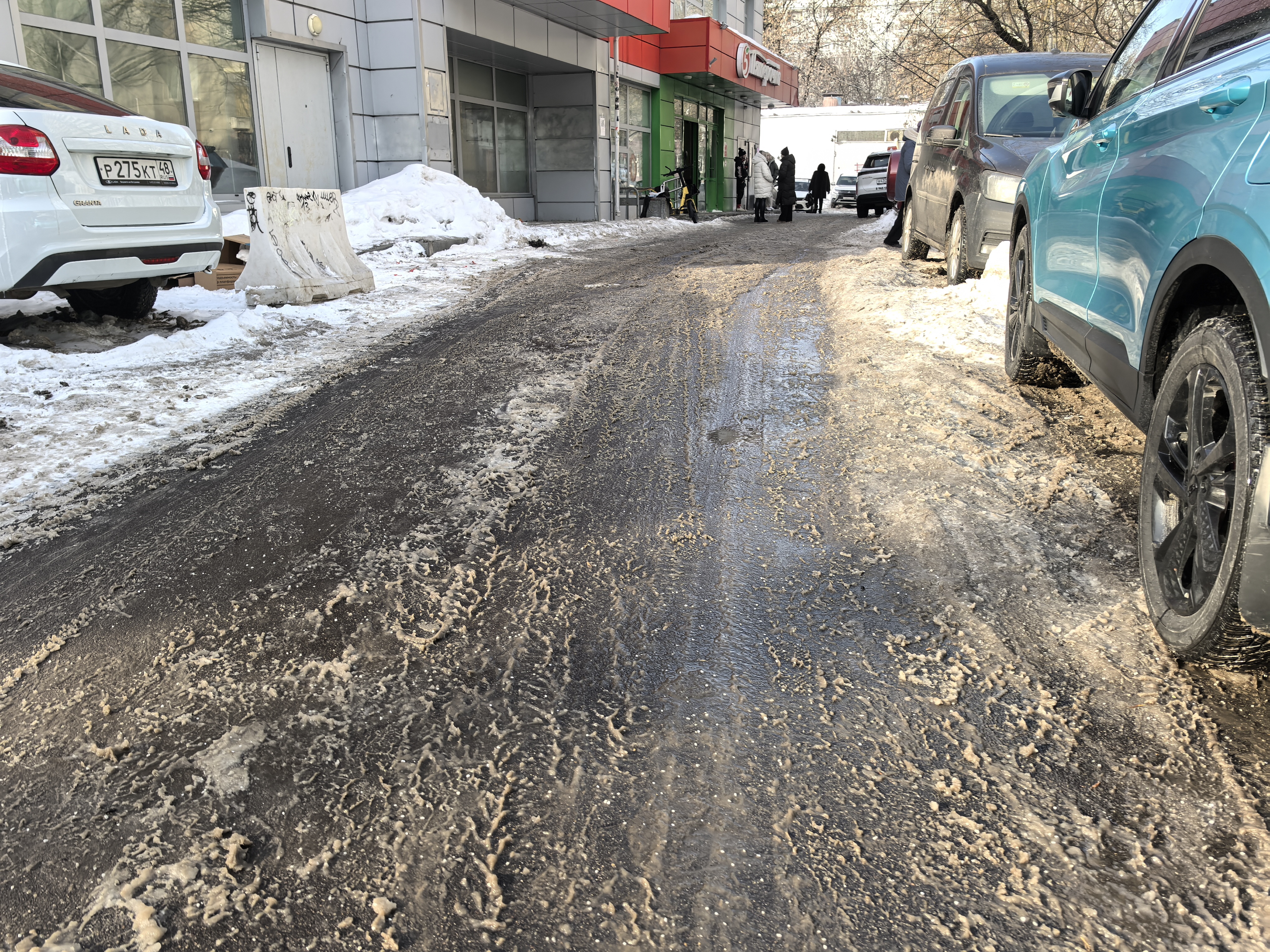 Дворовая территория с проезжей частью, покрытой грязью и снежной кашей. С обеих сторон припаркованы автомобили, тротуары отсутствуют или заблокированы снегом.
