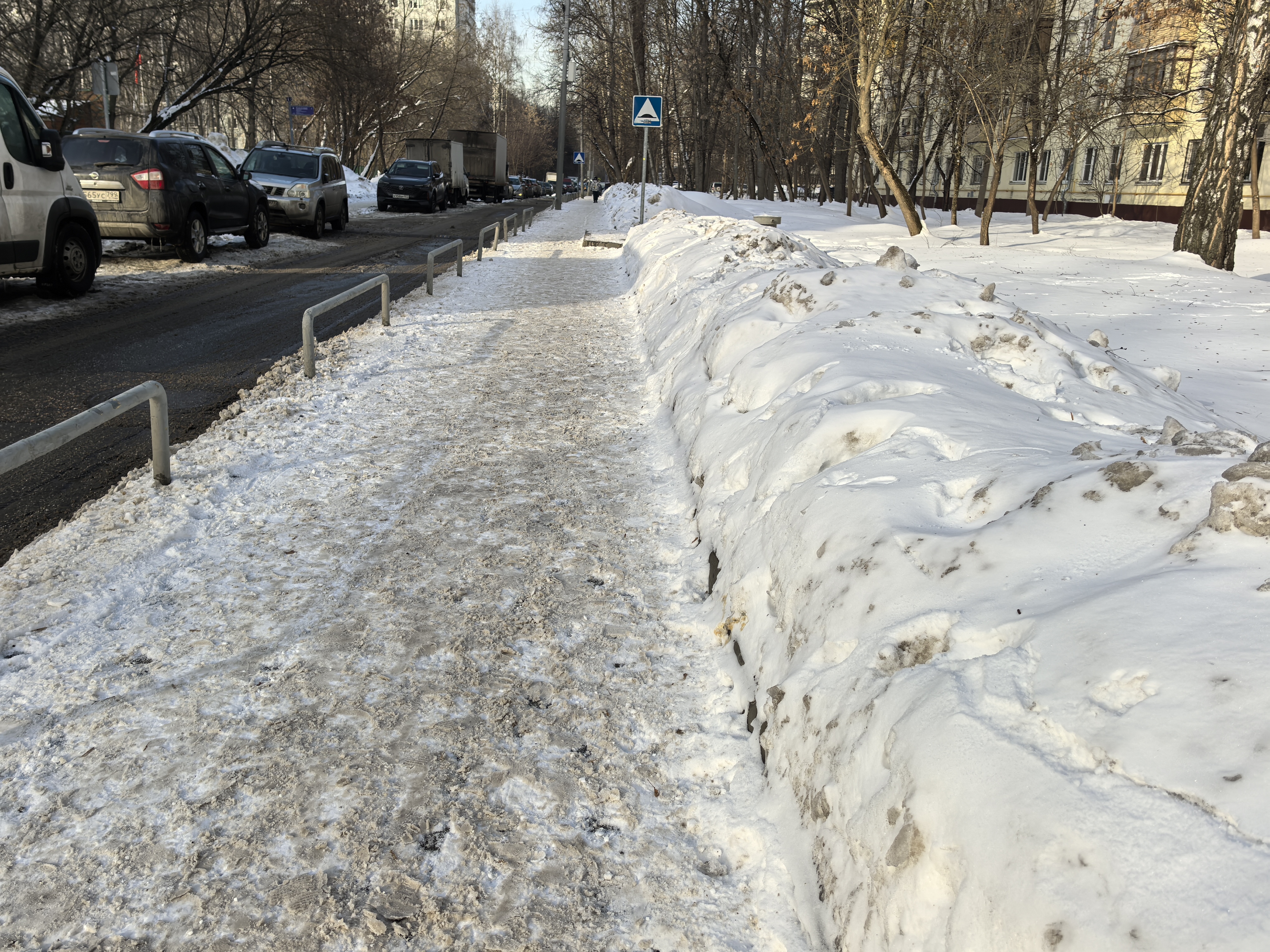 Участок городской дороги с тротуаром, покрытым снегом и ледяной коркой. Пешеходный переход отсутствует, но виден знак «Пешеходный переход» впереди. Тротуар отделен от дороги металлическими ограждениями.