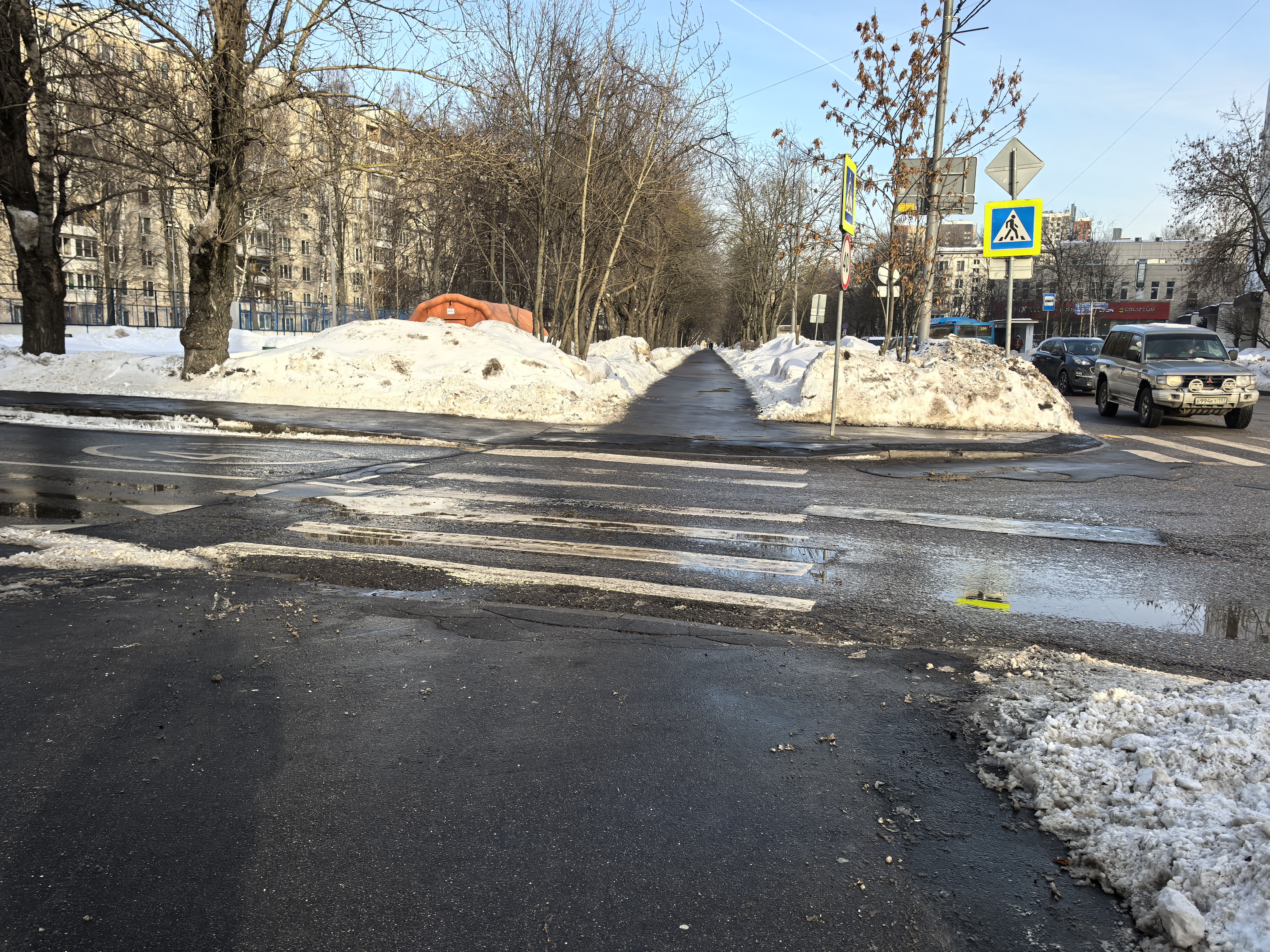 Участок проезжей части с пешеходным переходом (зебра), расположенный в жилой зоне. На дороге видны лужи и остатки снега, переход частично очищен, но разметка различима. Справа движется автомобиль, припаркованных машин у самого перехода нет.