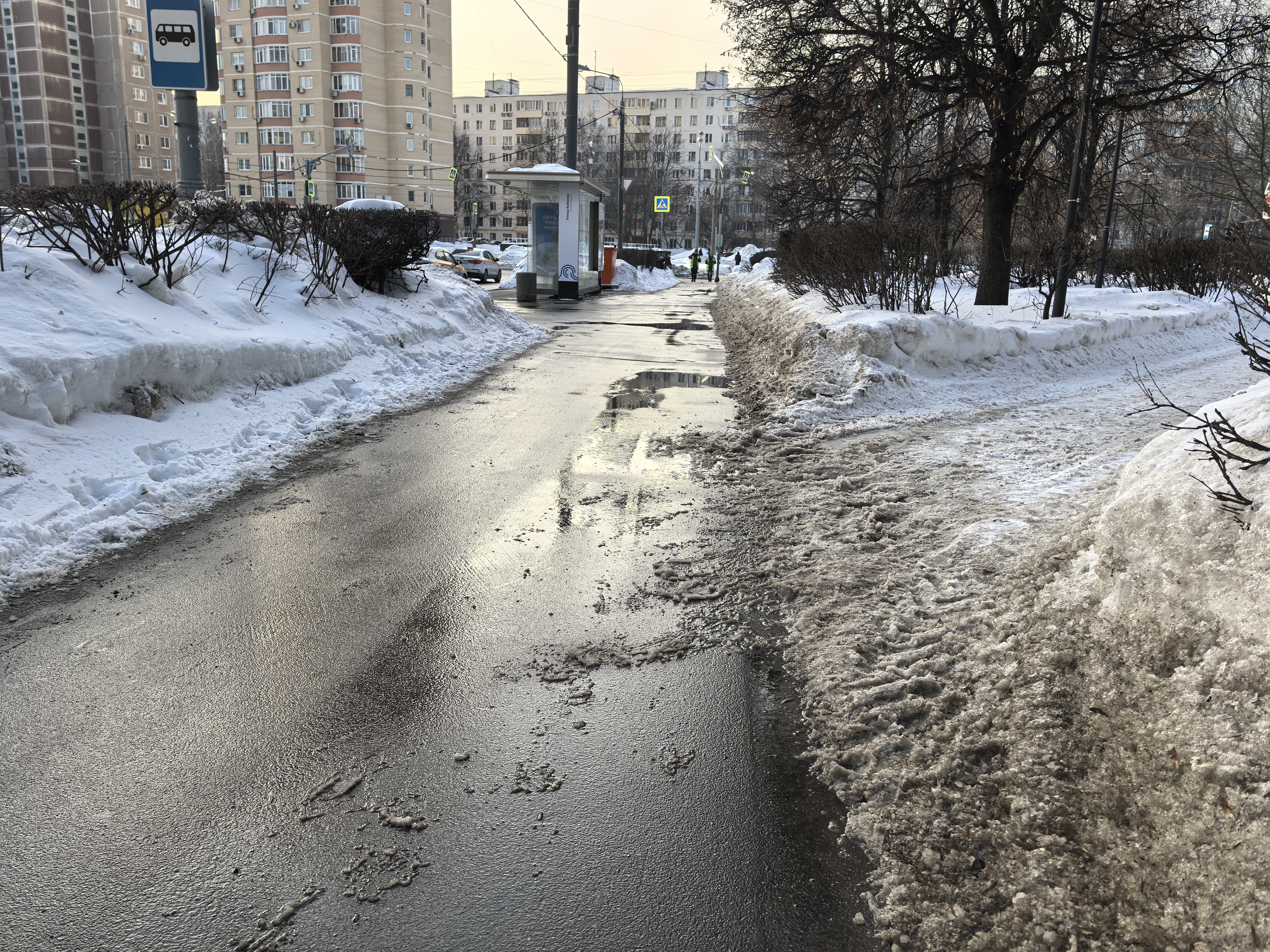 Участок дороги без пешеходного перехода, видна остановка общественного транспорта и тротуар. На проезжей части присутствуют лужи и грязь, тротуар очищен от снега, но имеет неровности.