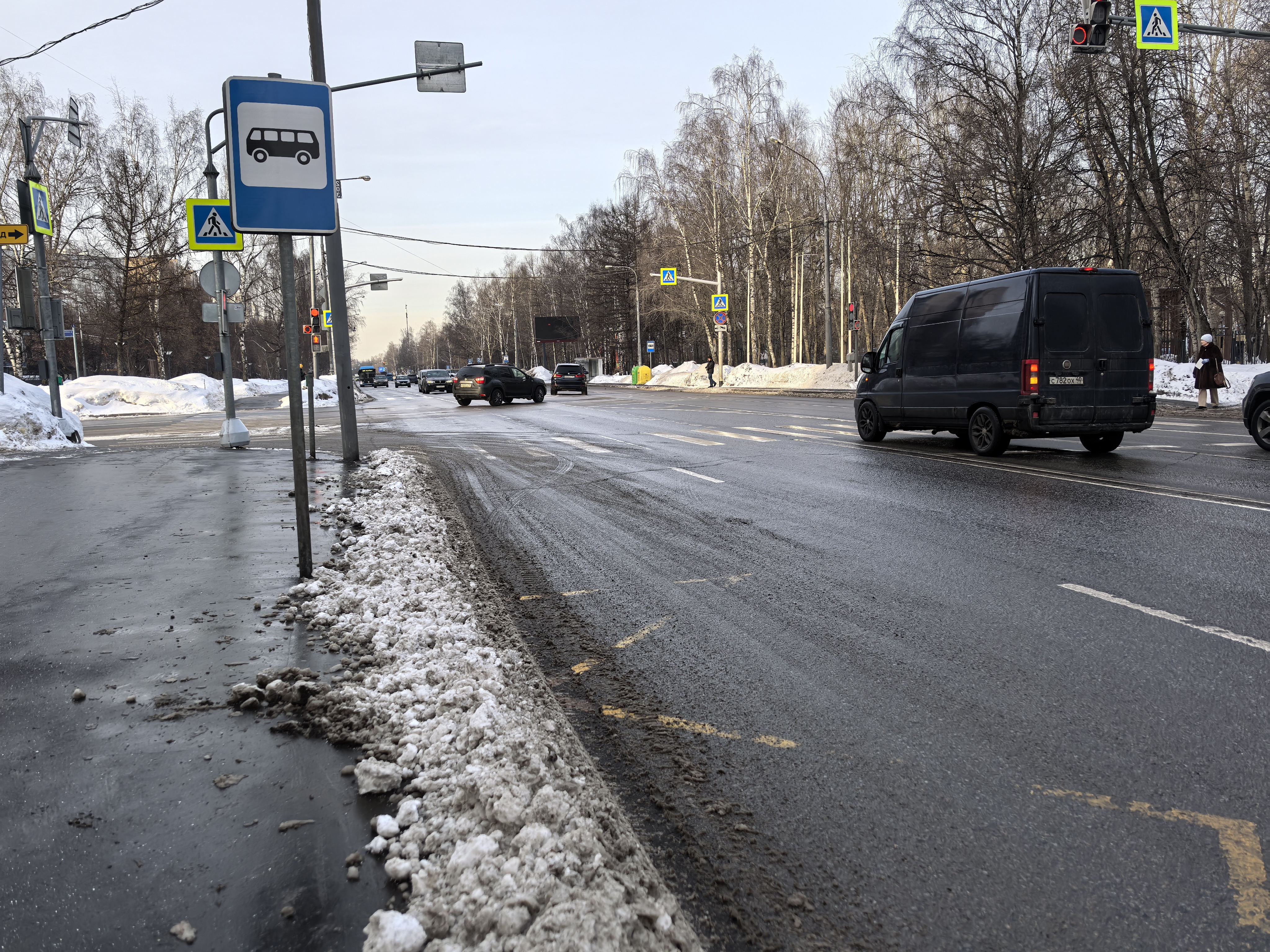 Участок проезжей части с пешеходным переходом, оборудованный светофором и знаками. На дороге видны движущиеся автомобили, припаркованных машин на переходе нет. Дорожное покрытие асфальтовое, мокрое, с заметными следами от снега и грязи по краям проезжей ч
