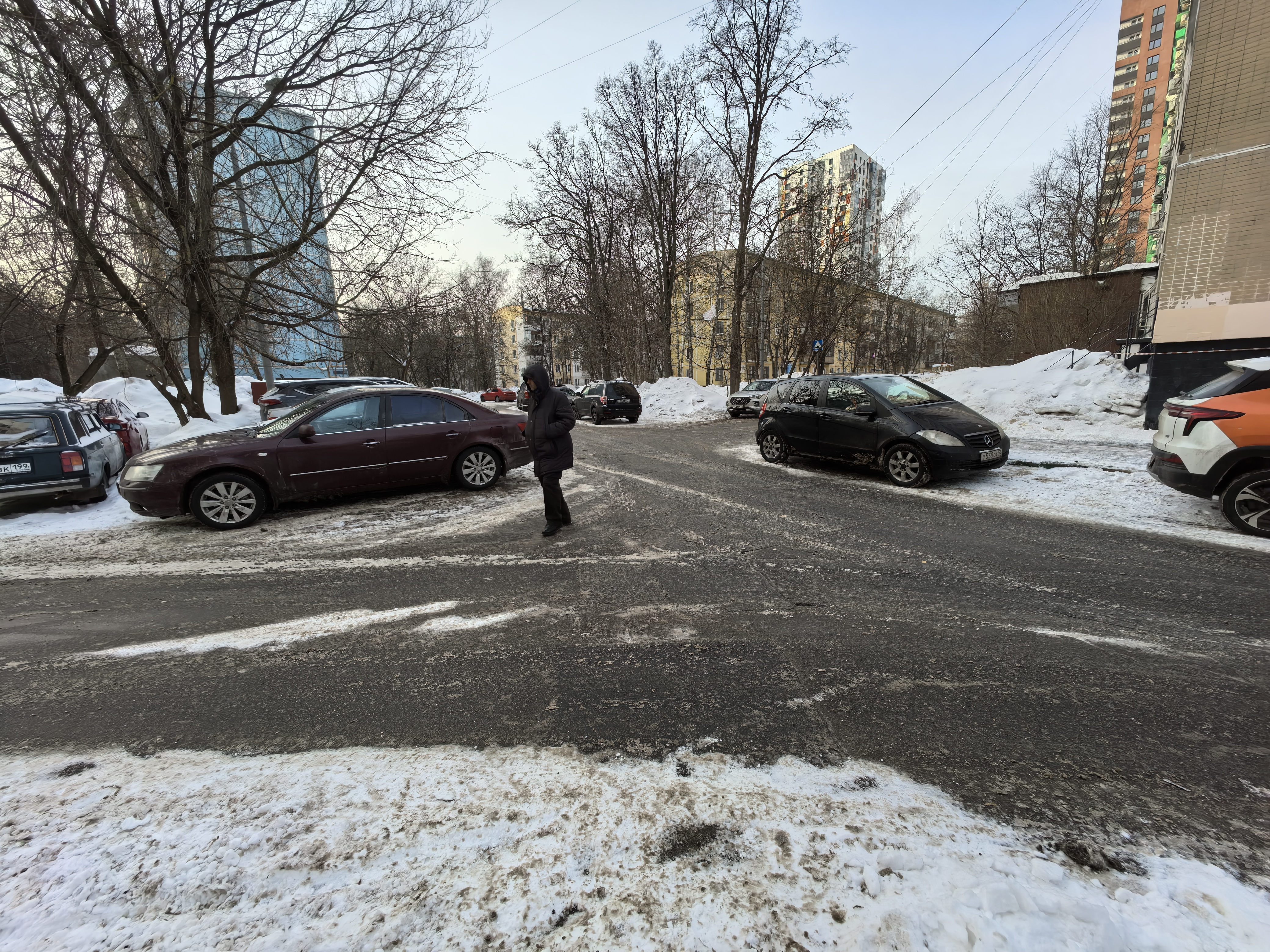 Дворовая территория с асфальтированной дорогой, окруженная жилыми домами. На дороге припаркованы автомобили, есть снег по краям.