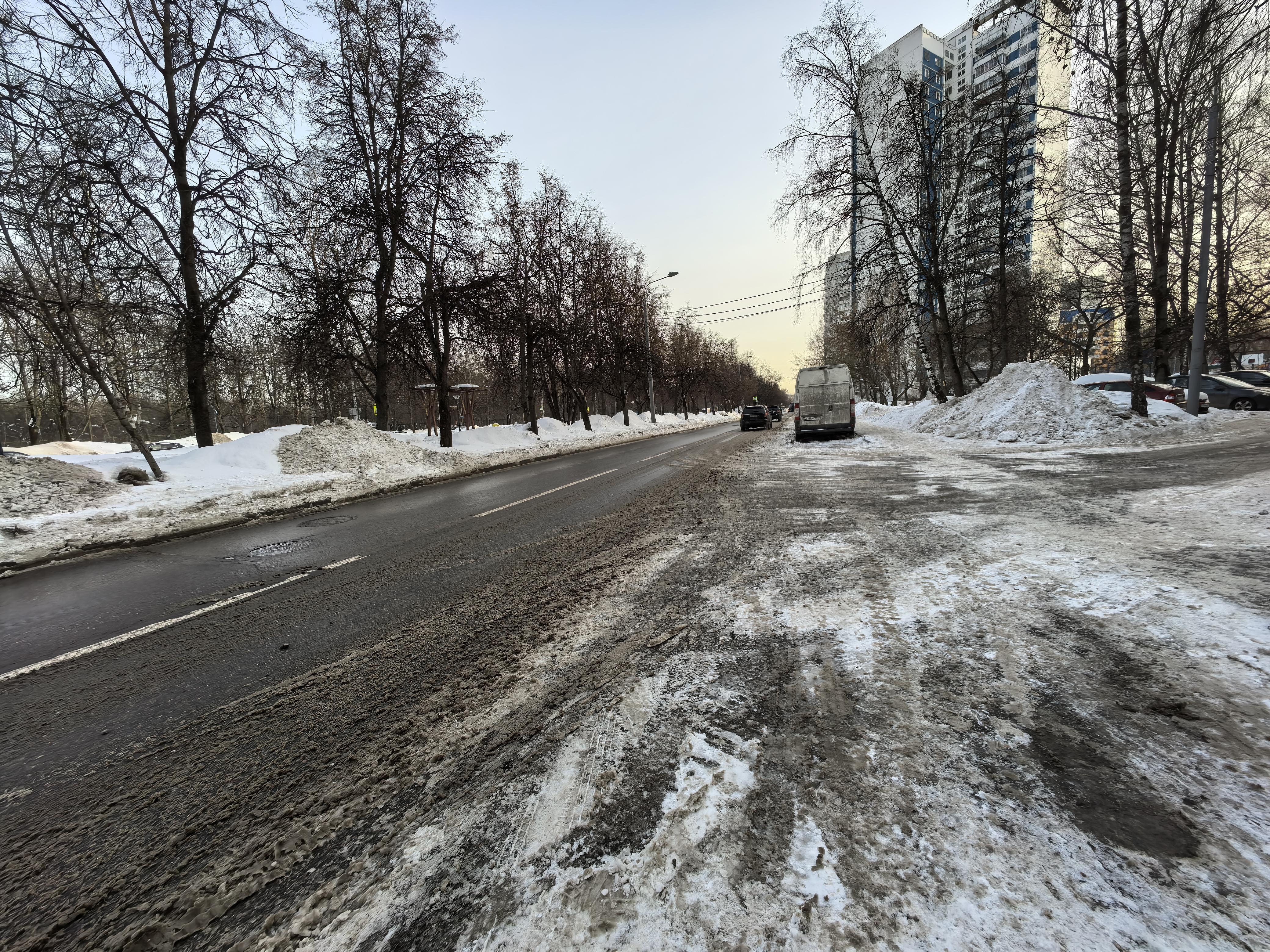 Проезжая часть без пешеходного перехода, виден тротуар слева, покрытый снегом, и припаркованные автомобили справа.