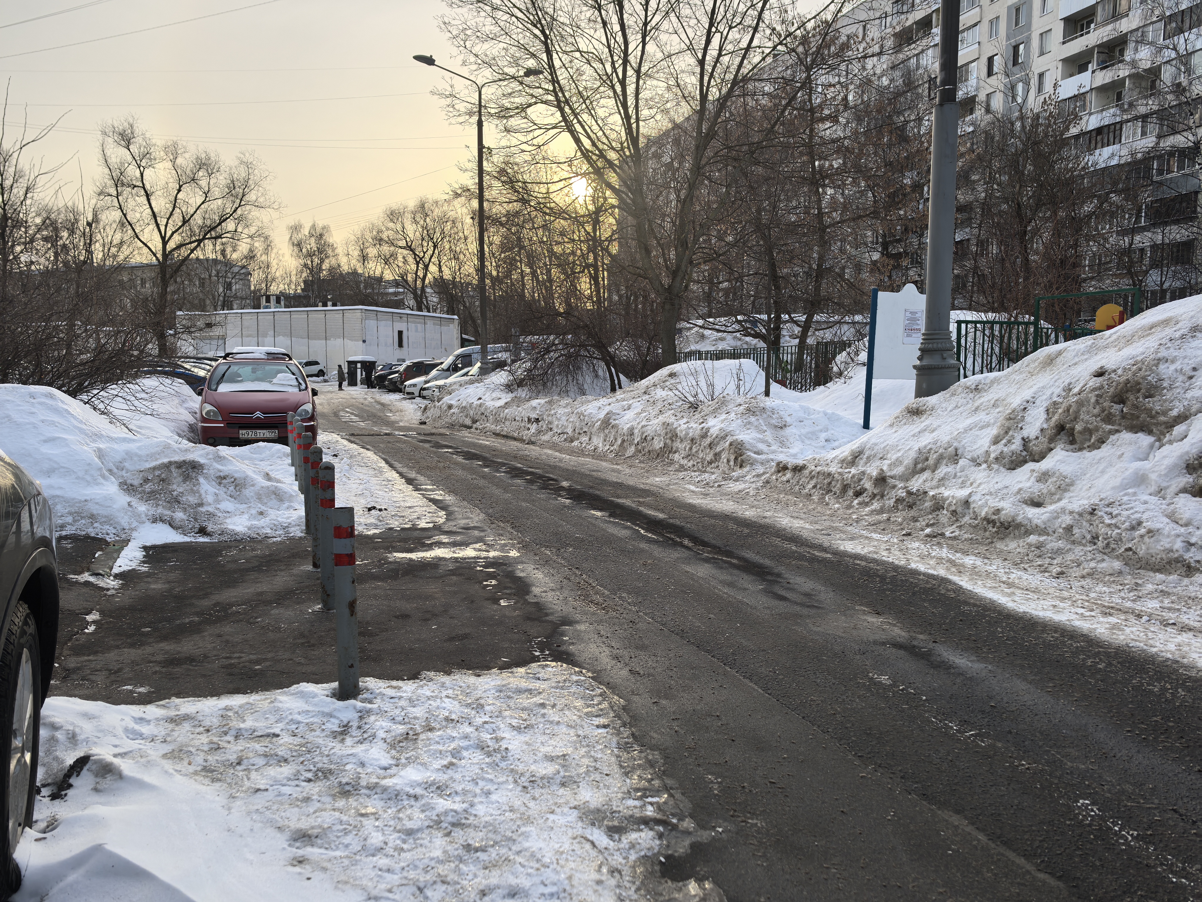 Дворовая территория с проезжей частью, окруженная жилыми домами и детской площадкой. Дорога узкая, с припаркованными автомобилями и сугробами по краям.