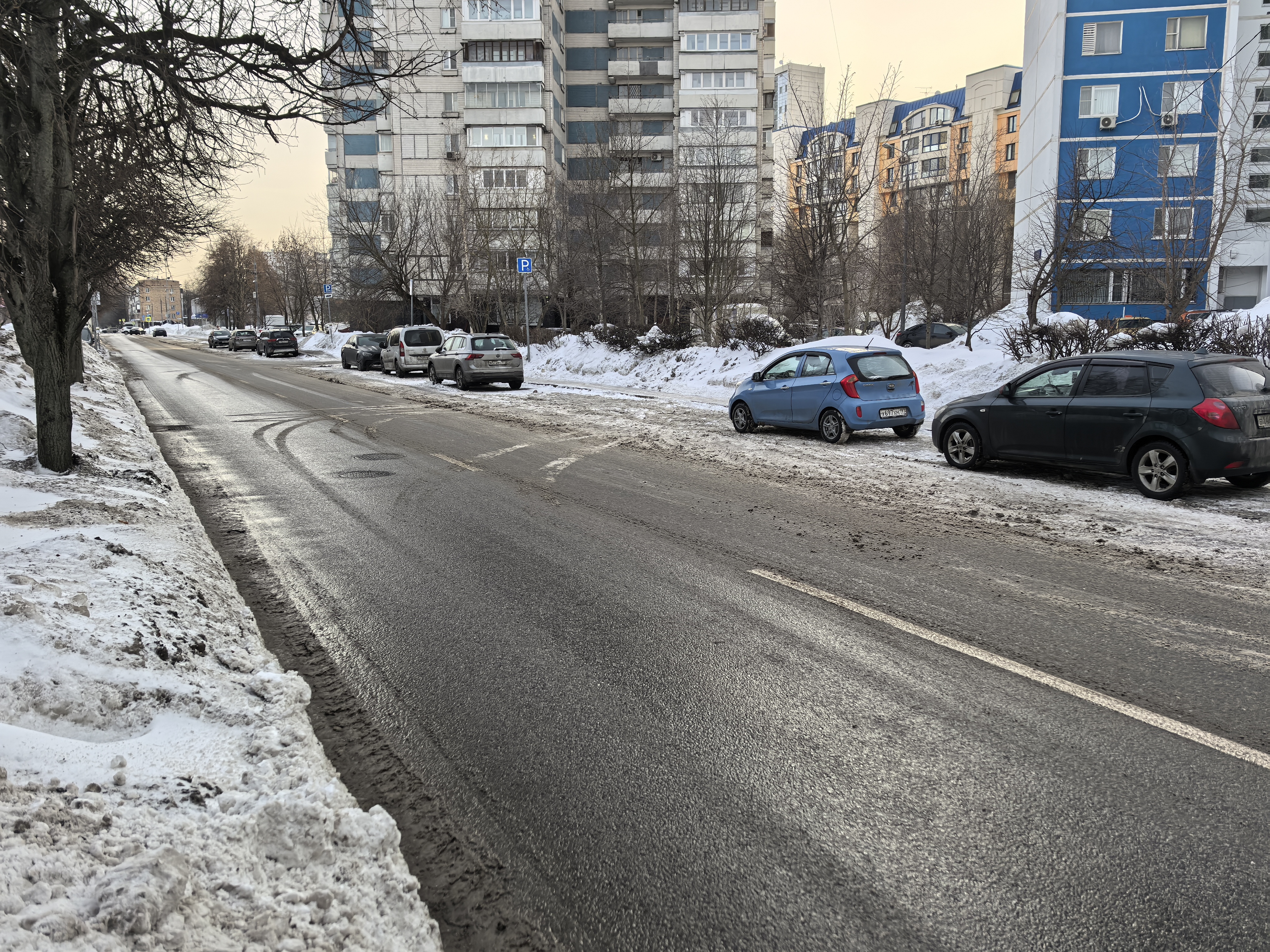 Городская улица с двусторонним движением, припаркованными автомобилями вдоль обочины, зимним покрытием и отсутствием пешеходного перехода на видимом участке.