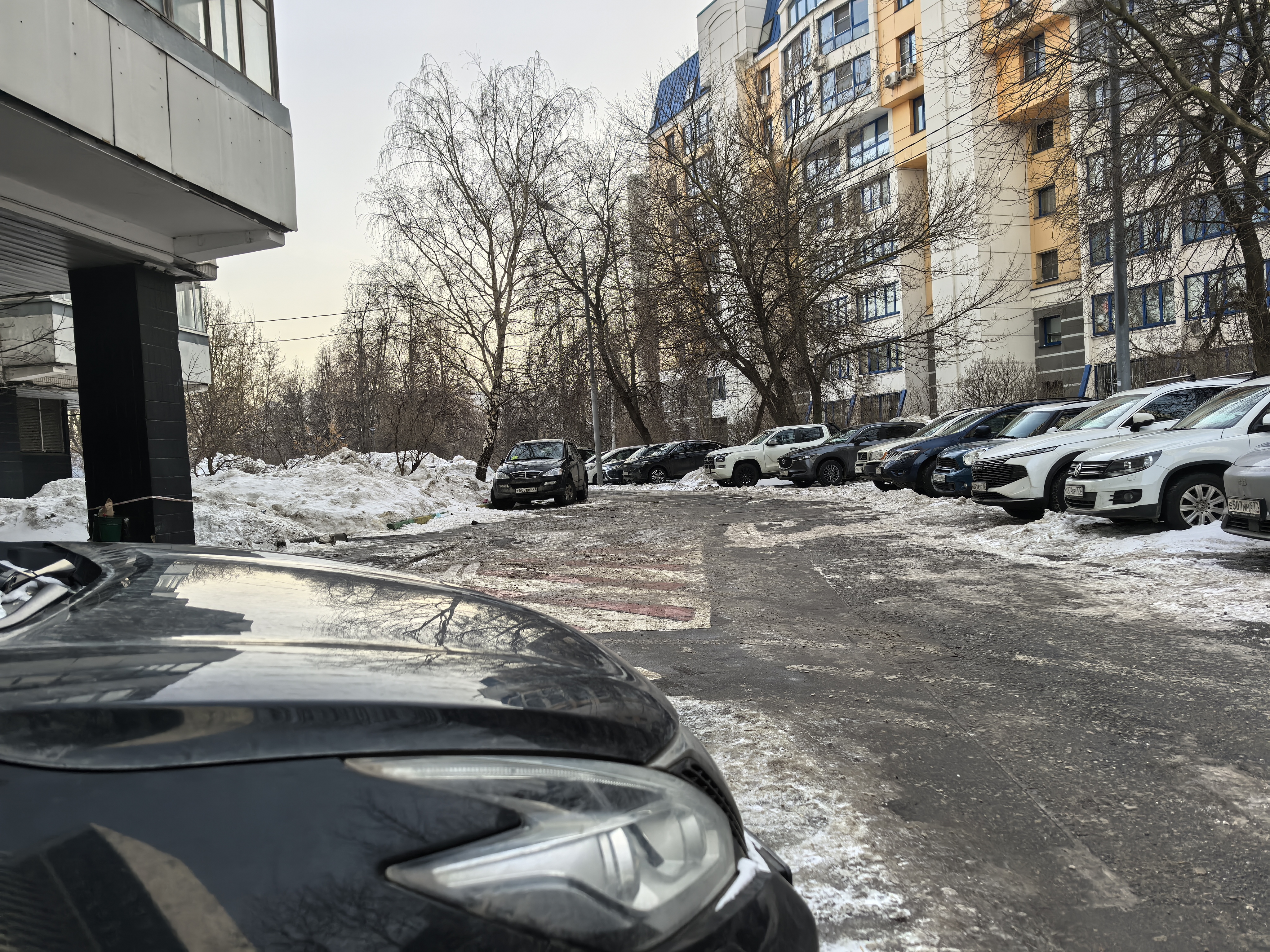 Дворовая территория с парковкой, покрытая снегом и льдом, с припаркованными автомобилями по обеим сторонам проезжей части