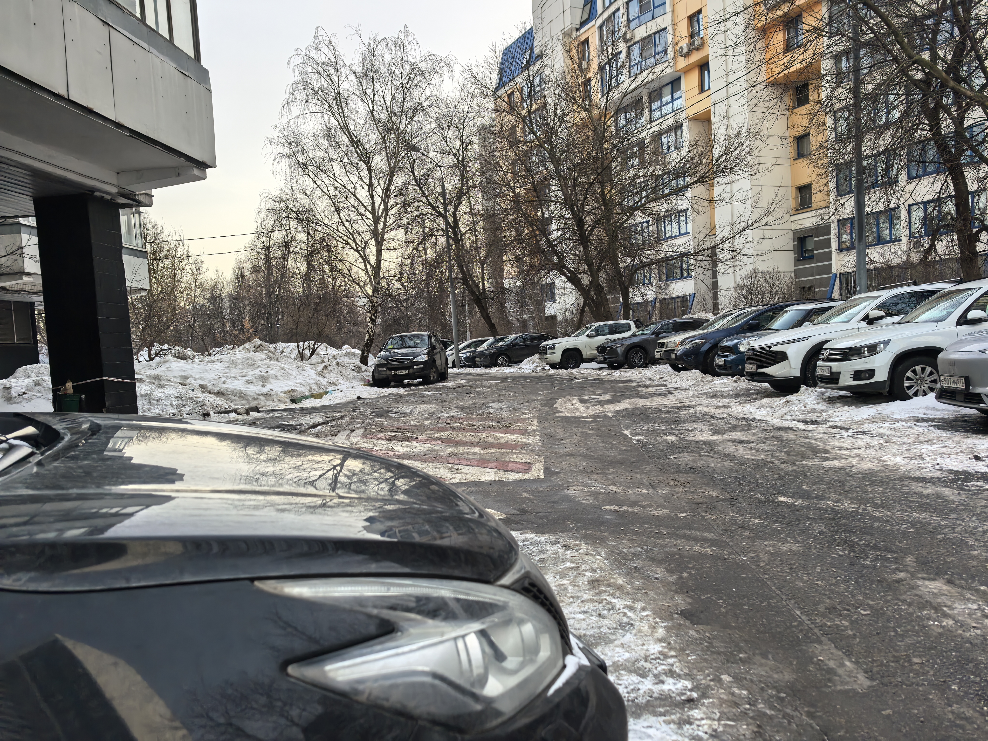 Участок дворовой территории с видимой пешеходной разметкой (зебра), окруженный припаркованными автомобилями и жилыми домами. Дорожное покрытие асфальтированное, частично покрыто снегом и наледью.