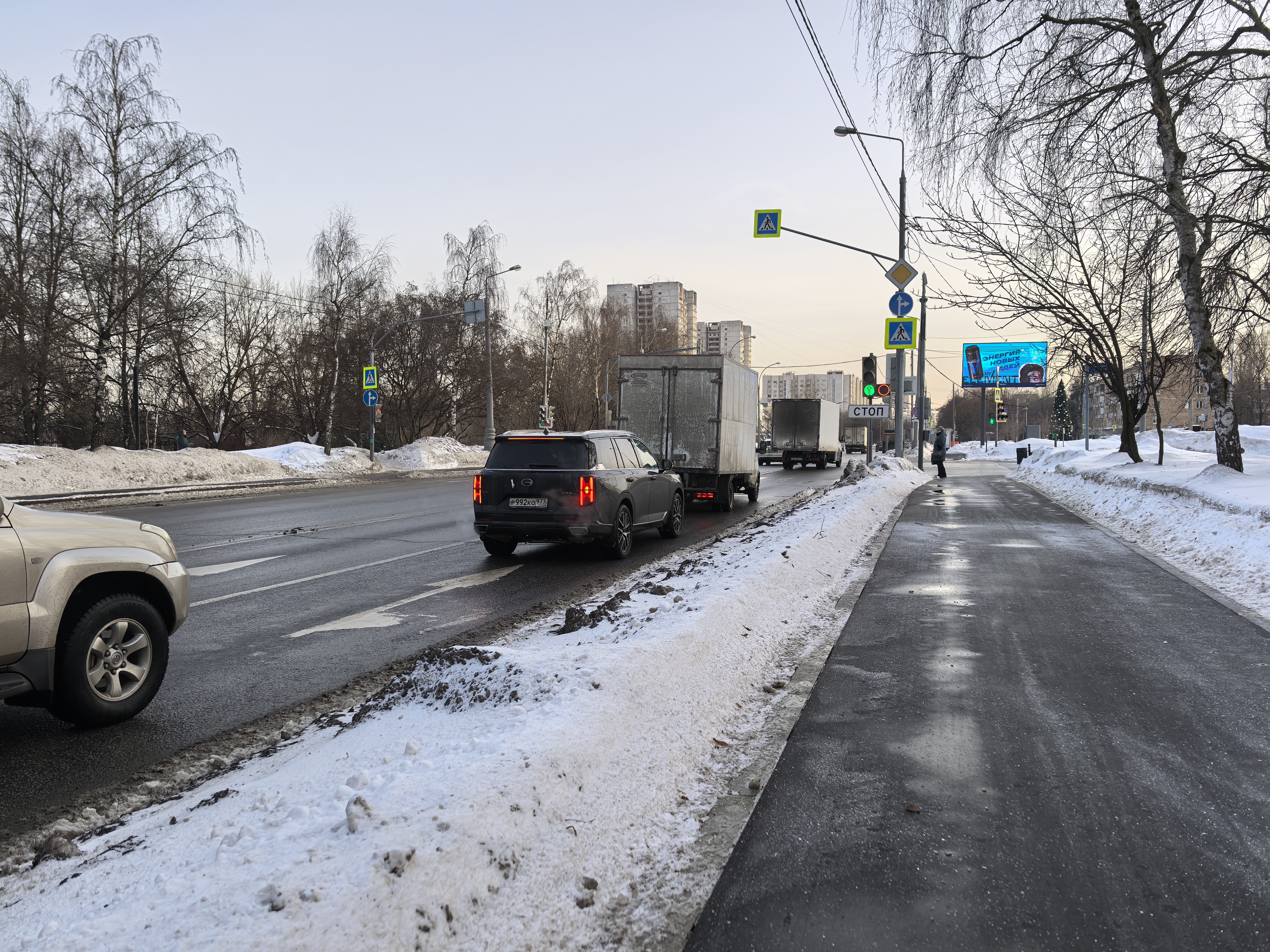 Участок проезжей части с пешеходным переходом, регулируемым светофором. На дороге присутствуют движущиеся транспортные средства, тротуар очищен от снега.