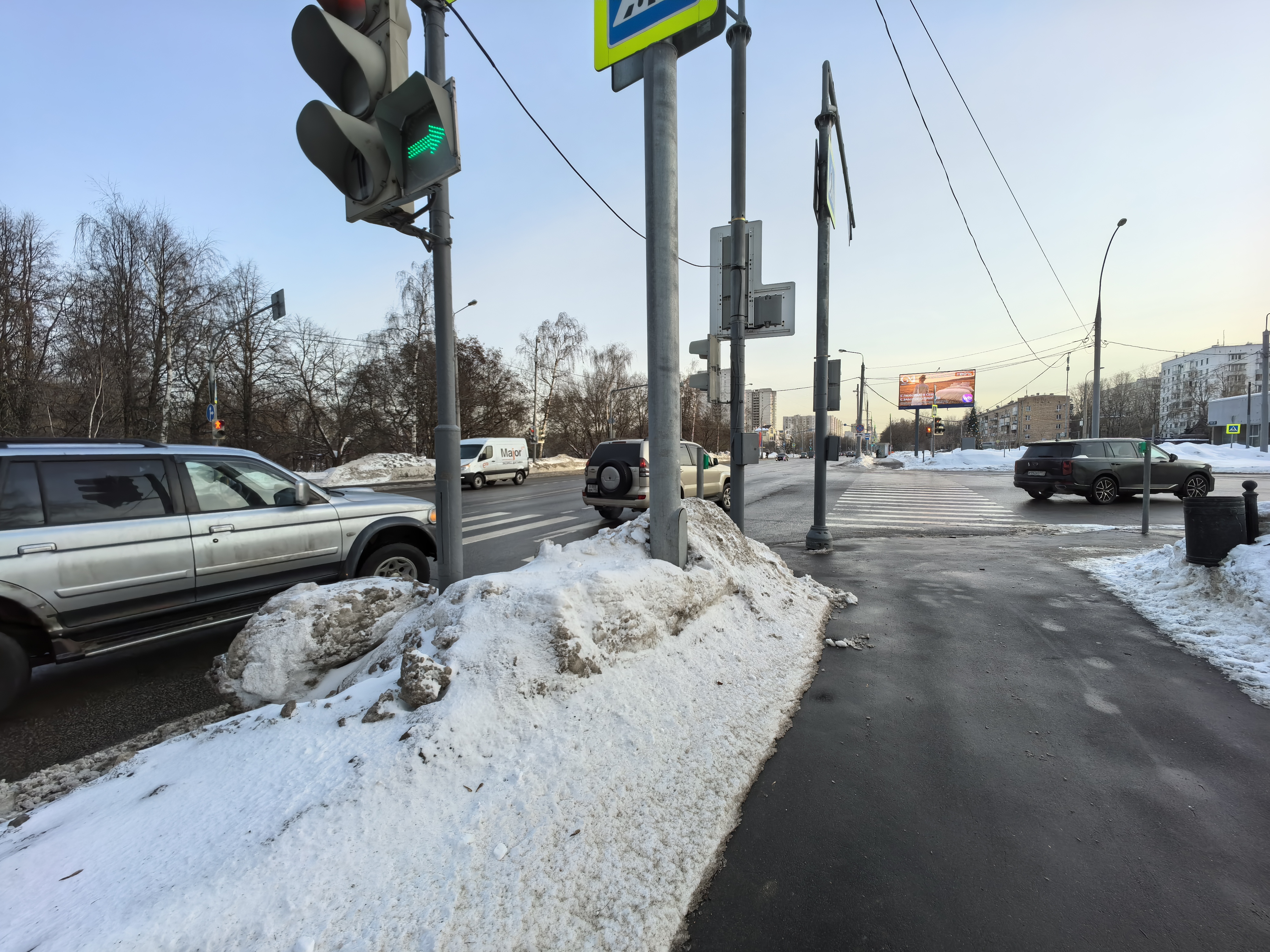 Перекресток с пешеходным переходом, оборудованный светофором. Дорожное покрытие чистое, но присутствуют снежные валы по краям проезжей части и тротуара.
