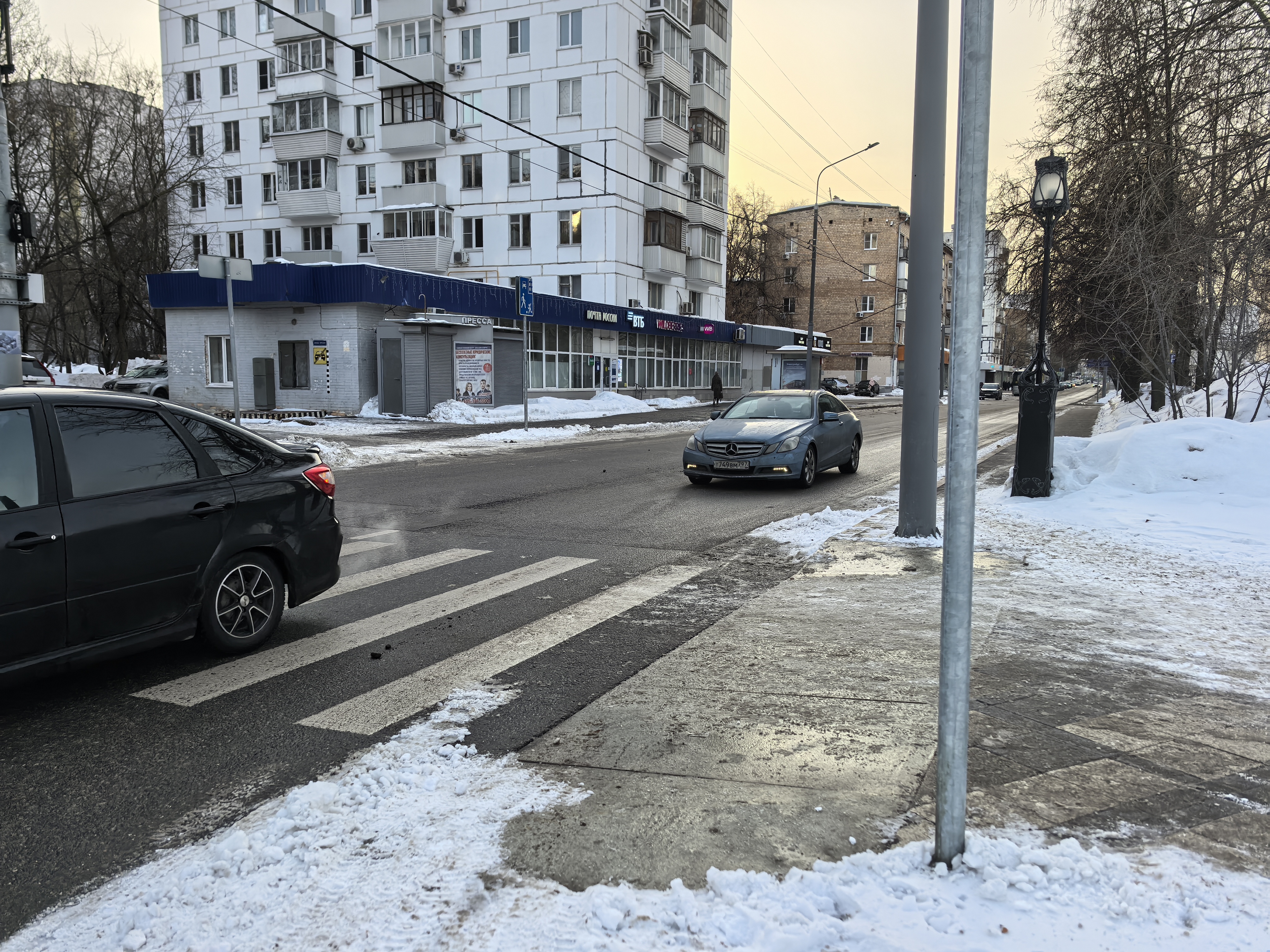 Городская улица с пешеходным переходом (зебра) и припаркованным автомобилем, частично перекрывающим обзор. На проезжей части виден движущийся автомобиль. Снег лежит на обочинах и тротуарах, но проезжая часть очищена.