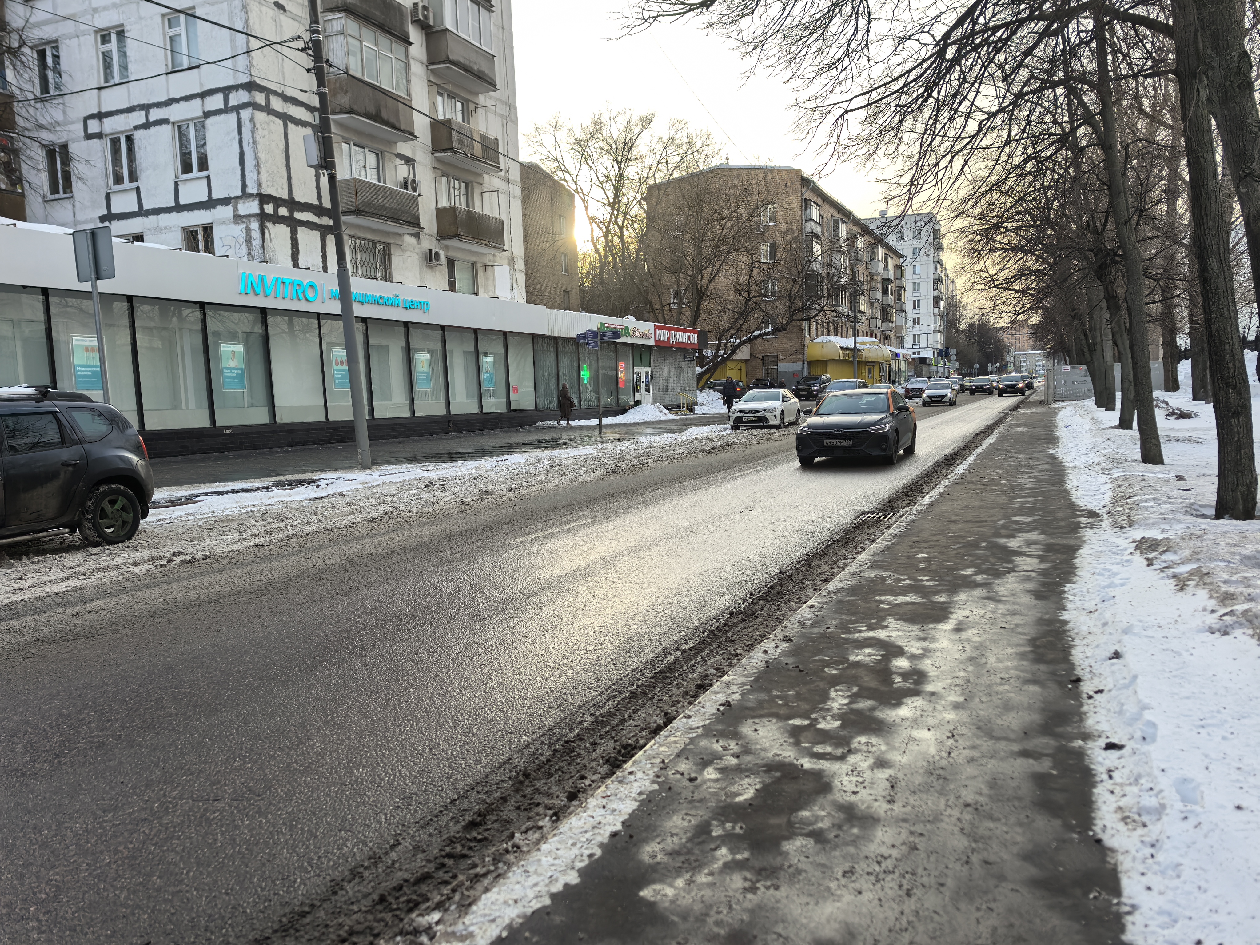 Участок городской улицы с проезжей частью и тротуаром, оборудованным бордюром. Пешеходный переход на видимом участке отсутствует, движение осуществляется по тротуару.