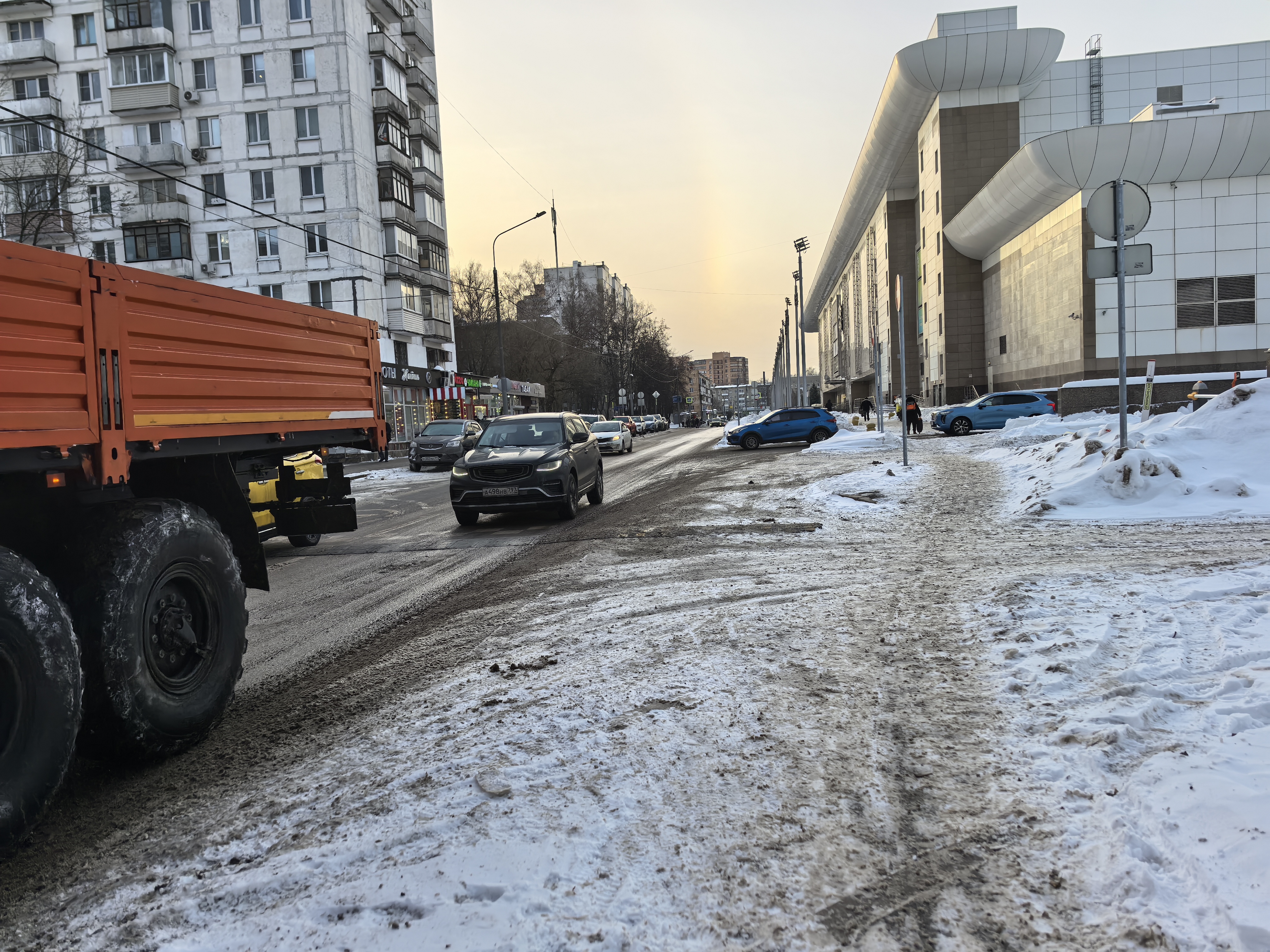 Участок городской дороги без обустроенного пешеходного перехода, покрытый снегом и грязью. Тротуары частично засыпаны снегом, проезжая часть занесена сугробами.