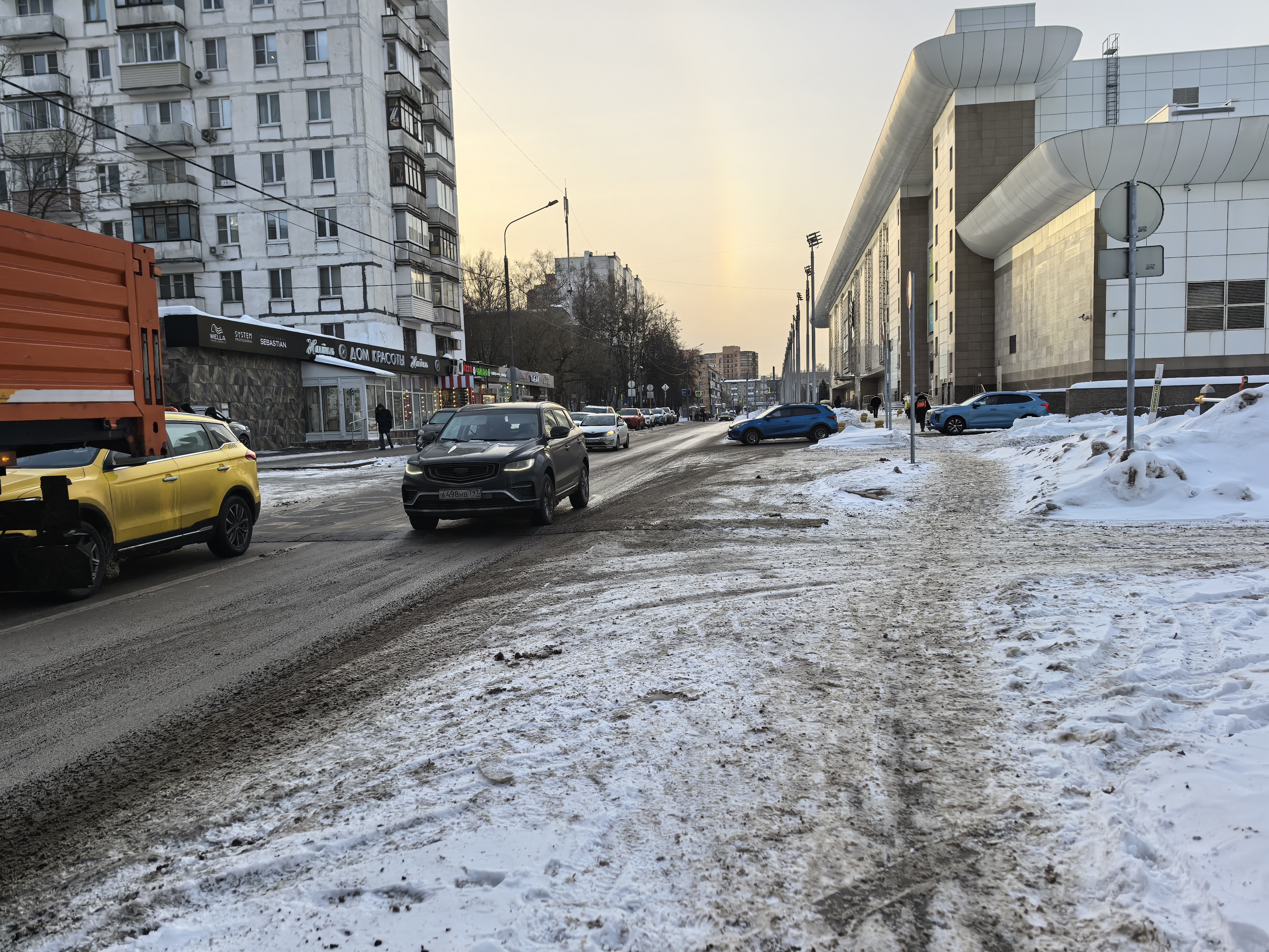 Городская улица с движением транспорта, пешеходный переход отсутствует. Тротуары частично покрыты снегом и грязью.