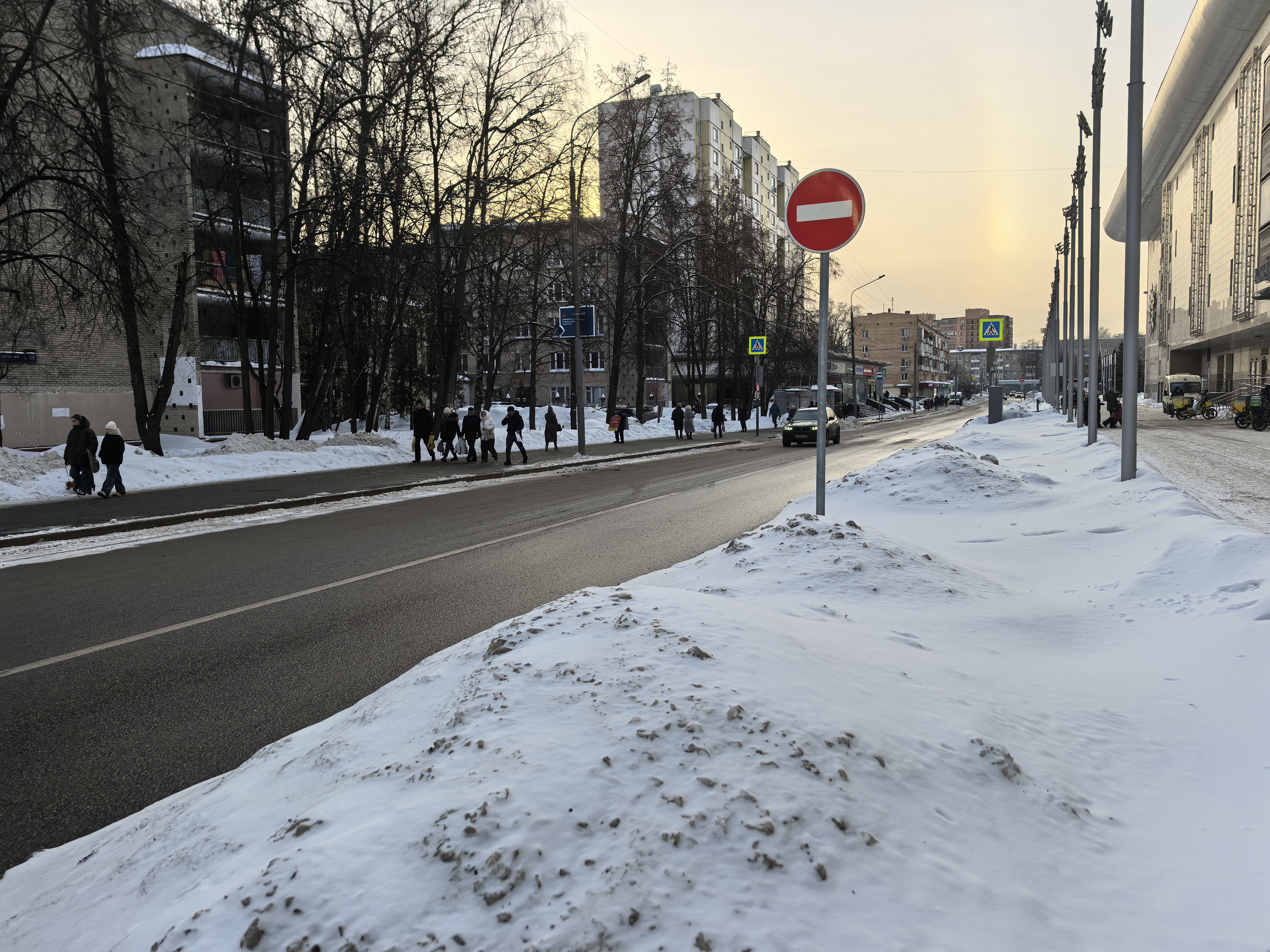 Городская улица с пешеходным переходом, видны пешеходы и дорожные знаки. Дорога покрыта снегом по краям, проезжая часть чистая.