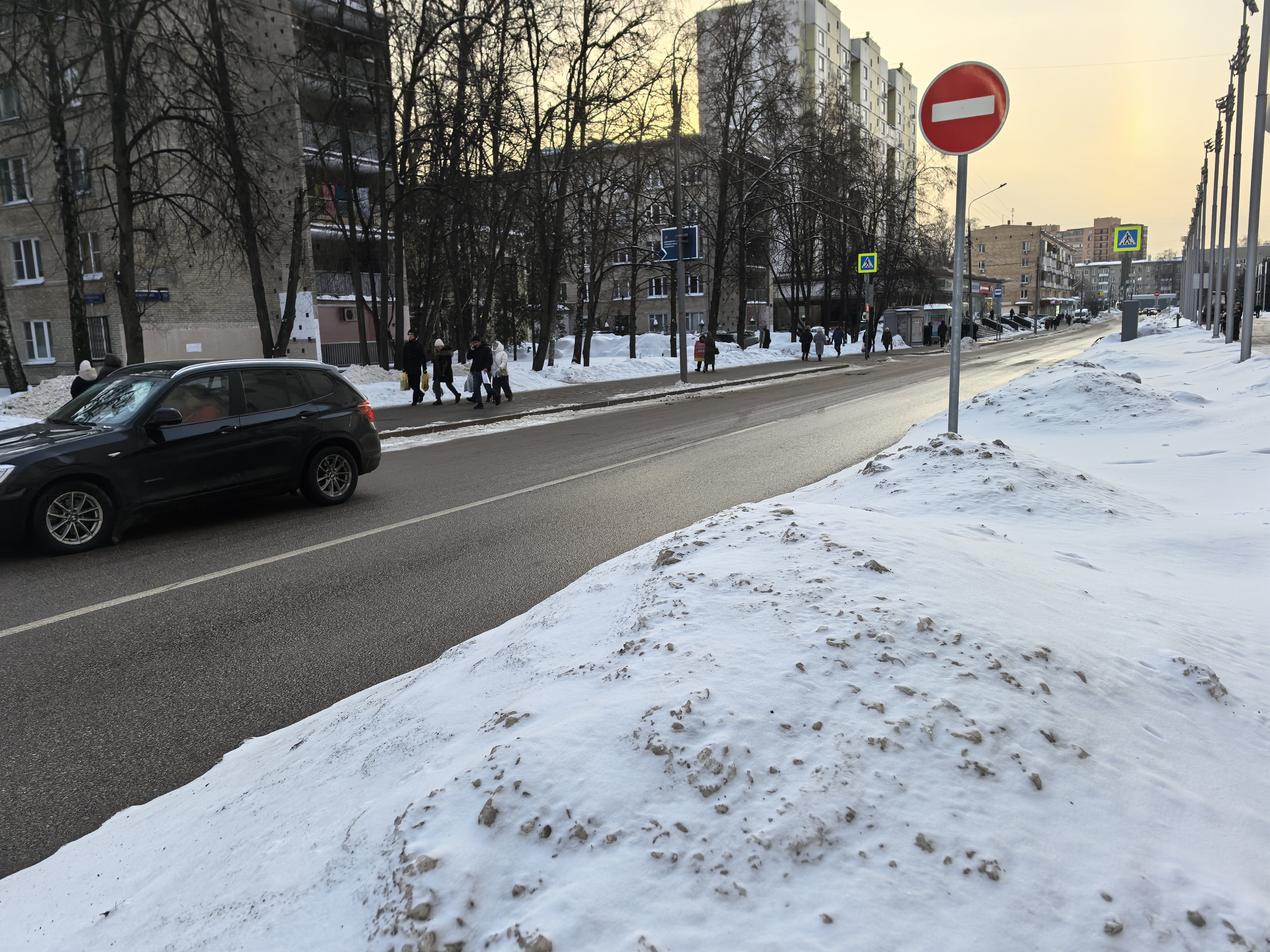 Городская улица с пешеходным переходом, оборудованным знаками и разметкой. На проезжей части виден движущийся автомобиль, на тротуаре — пешеходы. Зимний период, снег на обочинах.